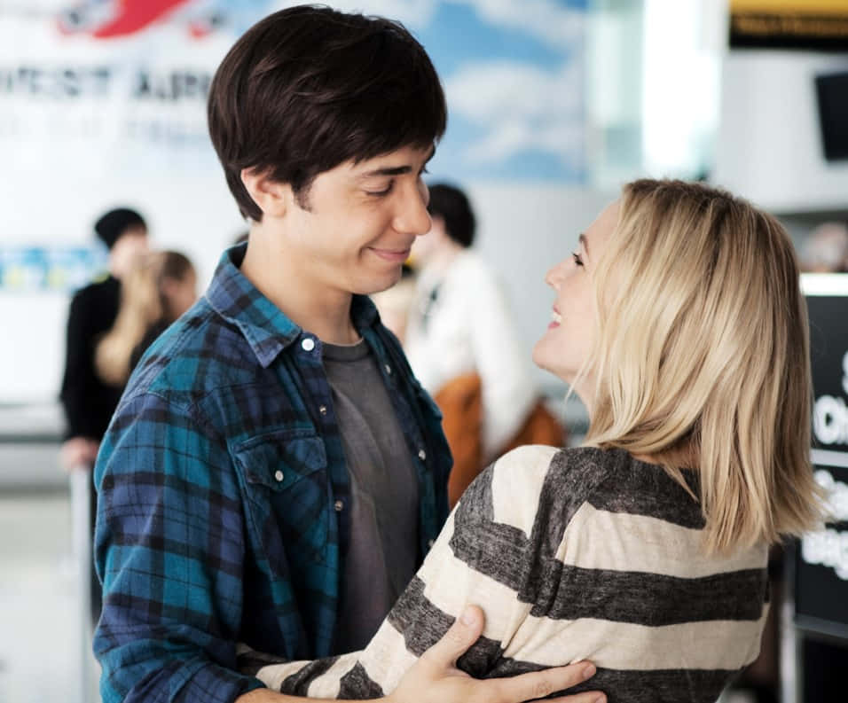 Justin Long 桌布 壁紙 — 一對微笑的年輕情侶在熙來攘往的機場客運大樓中熱情擁抱，背景有旅客和指示牌。