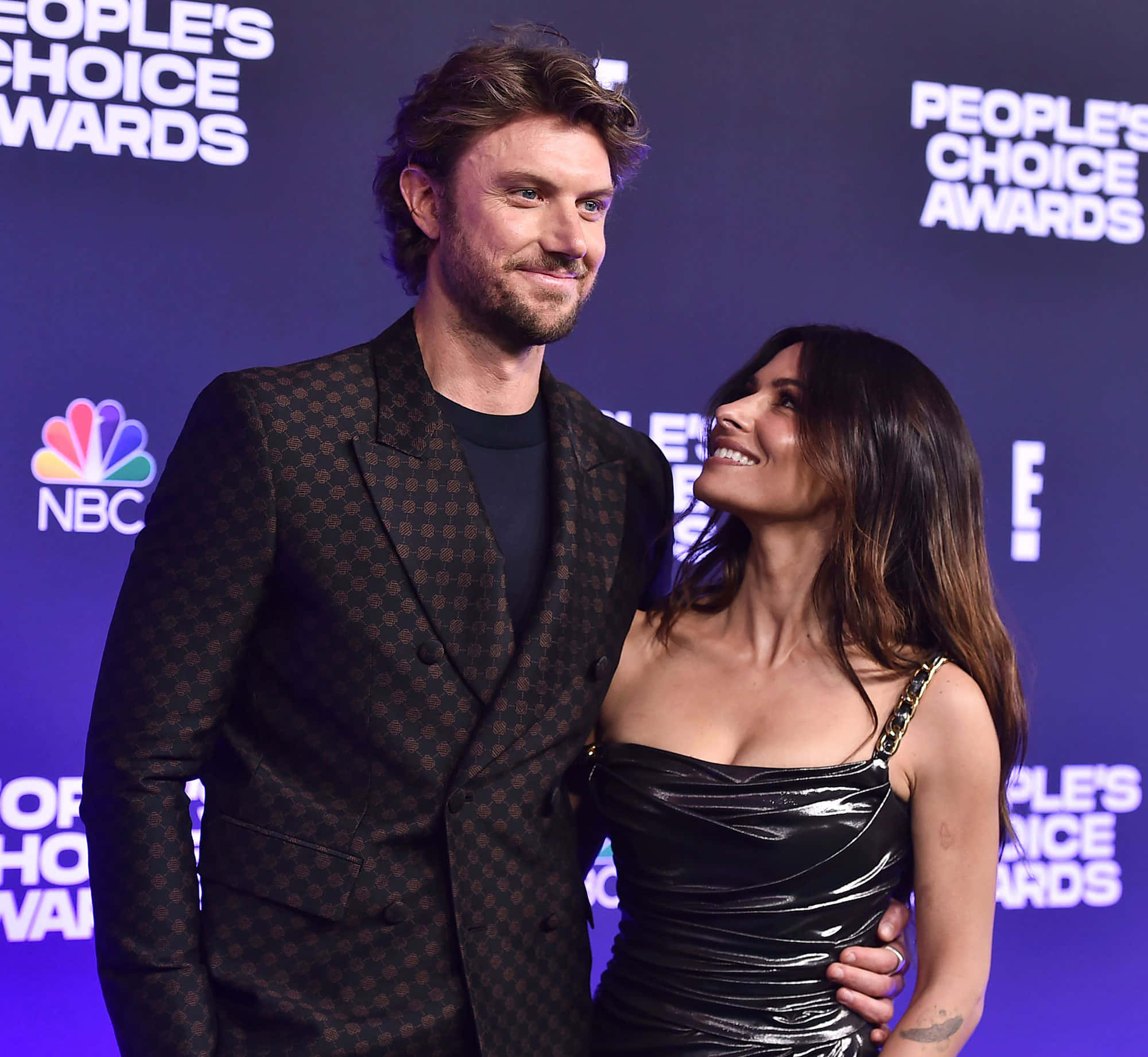 sarah shahi 桌布 壁紙 — 一對微笑的情侶在人民選擇獎的紫色品牌背景前合照。