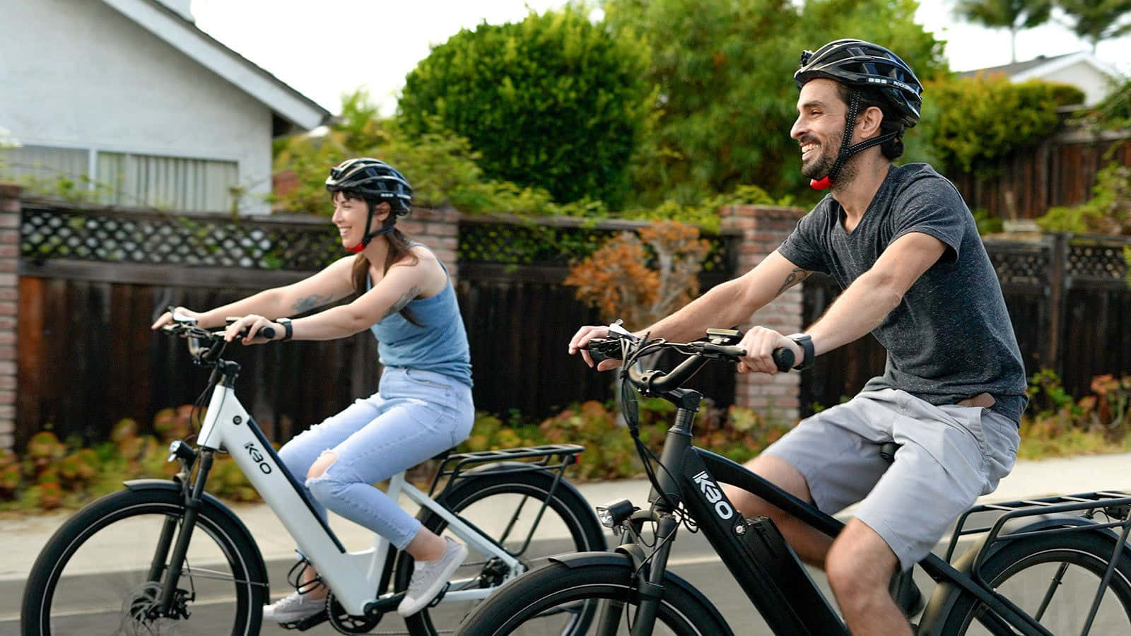 Electric Bike 桌布 壁紙 — 一對微笑的情侶在綠樹成蔭、木柵欄環繞的郊區街道上騎電動單車。