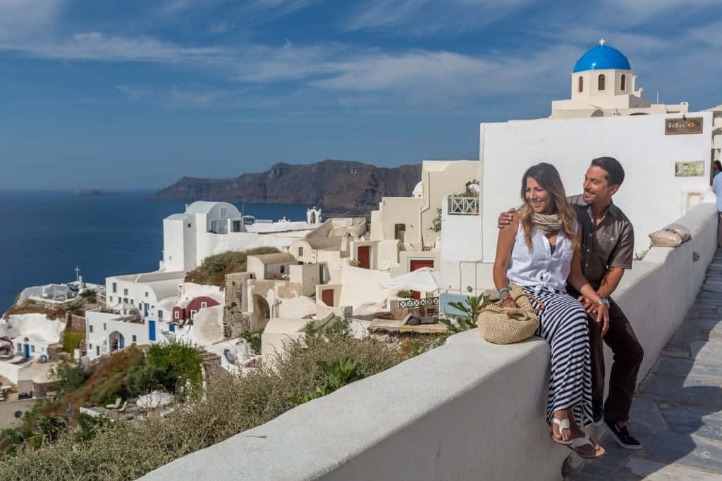 Greek Island 桌布 壁紙 — 一對微笑的情侶坐在白色石牆上，俯瞰著奧伊亞聖托里尼標誌性的藍頂教堂和白色建築，背景是明亮的藍天。