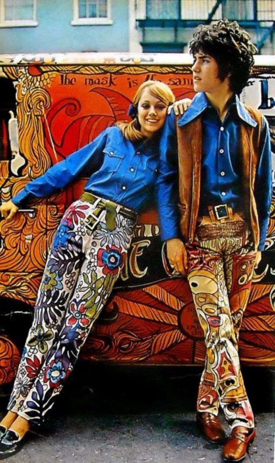 70年代gif 桌布 壁紙 — 一對微笑的情侶穿著迷幻的1970年代服裝，在一輛塗有鮮豔漩渦圖案和文字的貨車旁擺姿勢。