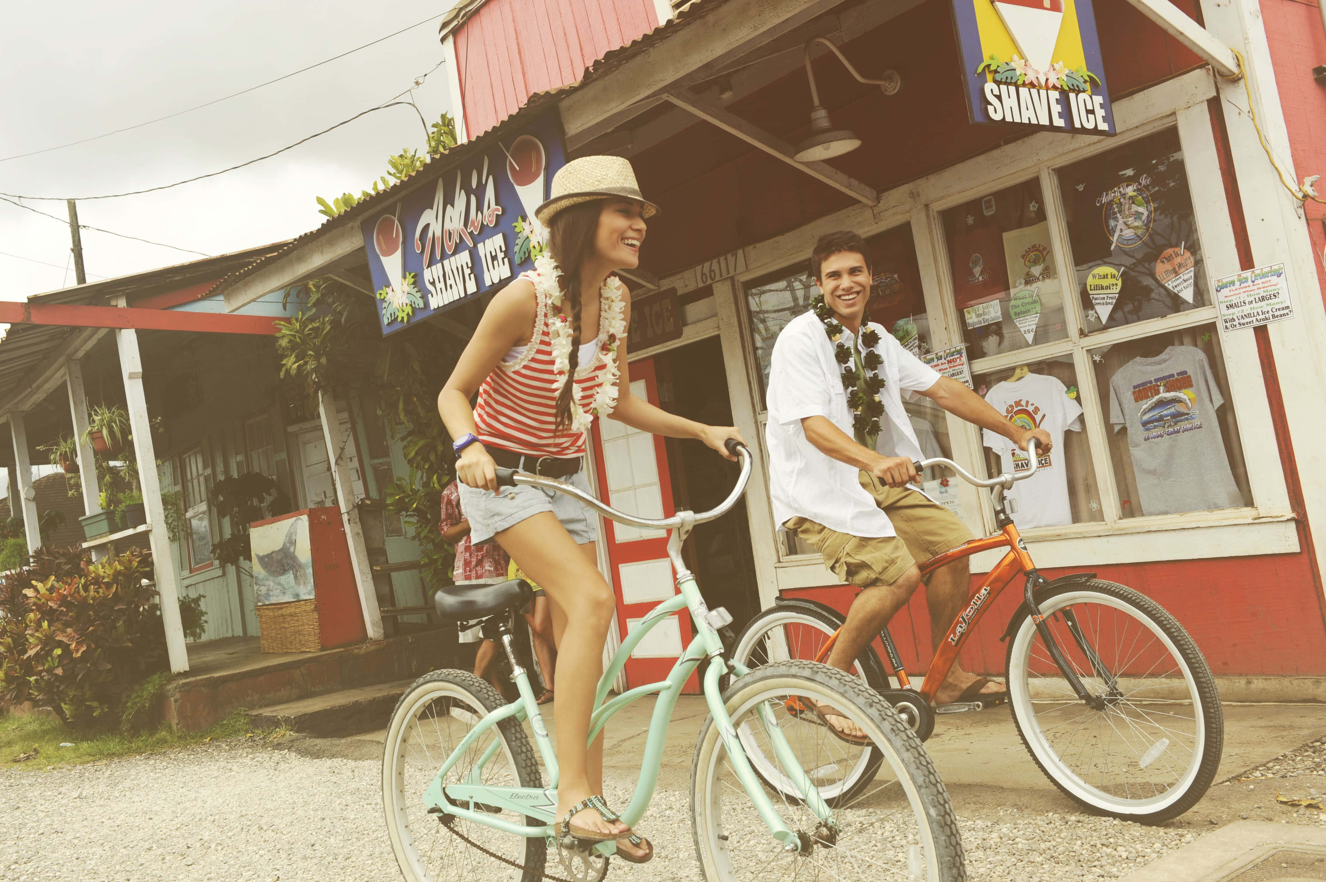 Beach Cruiser 桌布 壁紙 — 一對微笑的情侶踩單車經過陽光明媚的熱帶街道上一間色彩繽紛的刨冰店。