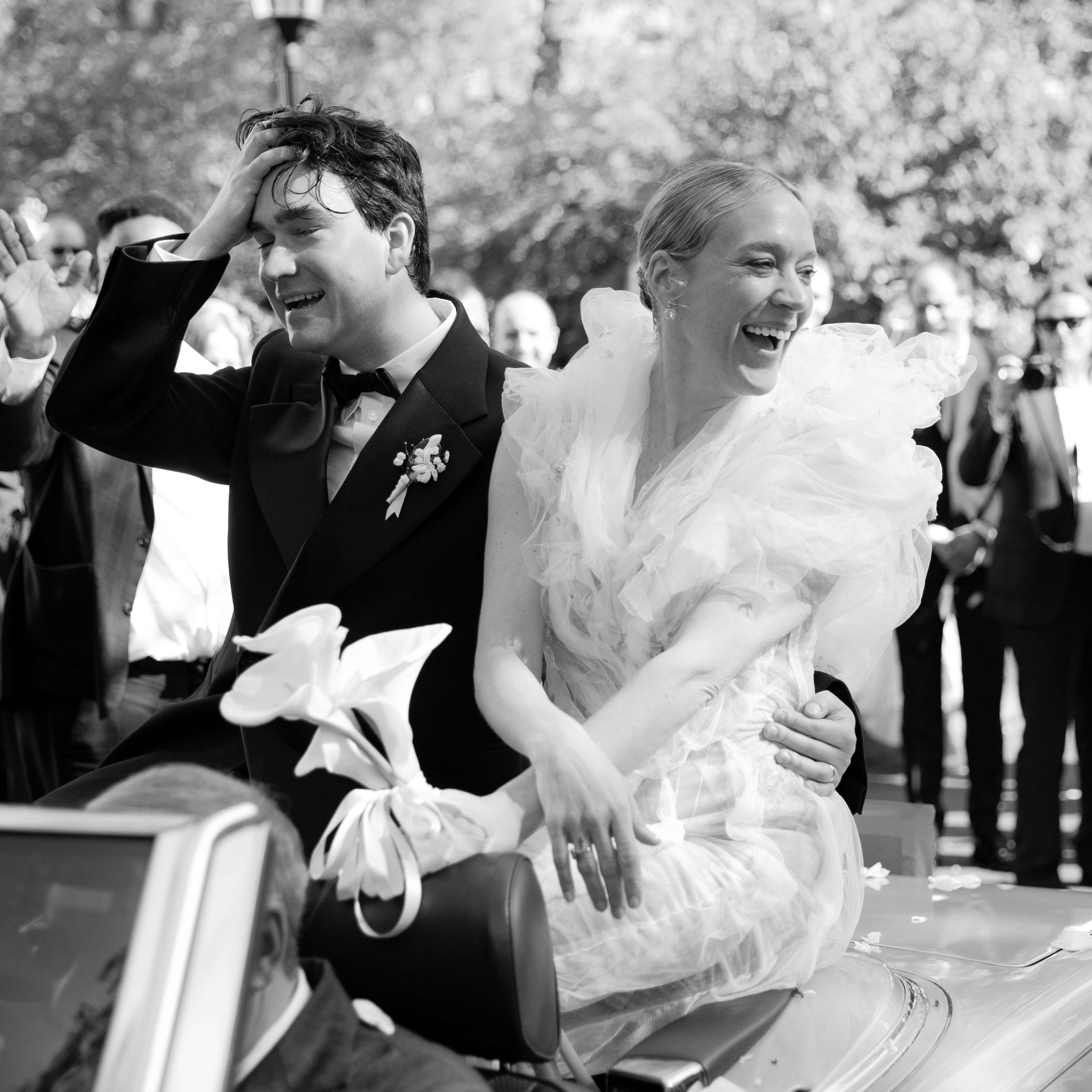 Chloë Sevigny 桌布 壁紙 — 一對快樂的新娘和新郎在黑白畫面中乘坐復古車穿過人群，開懷大笑。