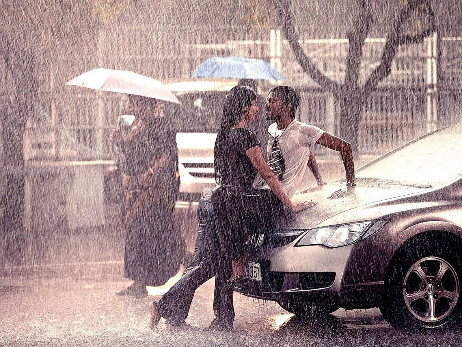 情侶 桌布 壁紙 — 一對情侶在傾盆大雨中靠著一輛銀色汽車分享親密時刻，附近另一個人撐著雨傘。
