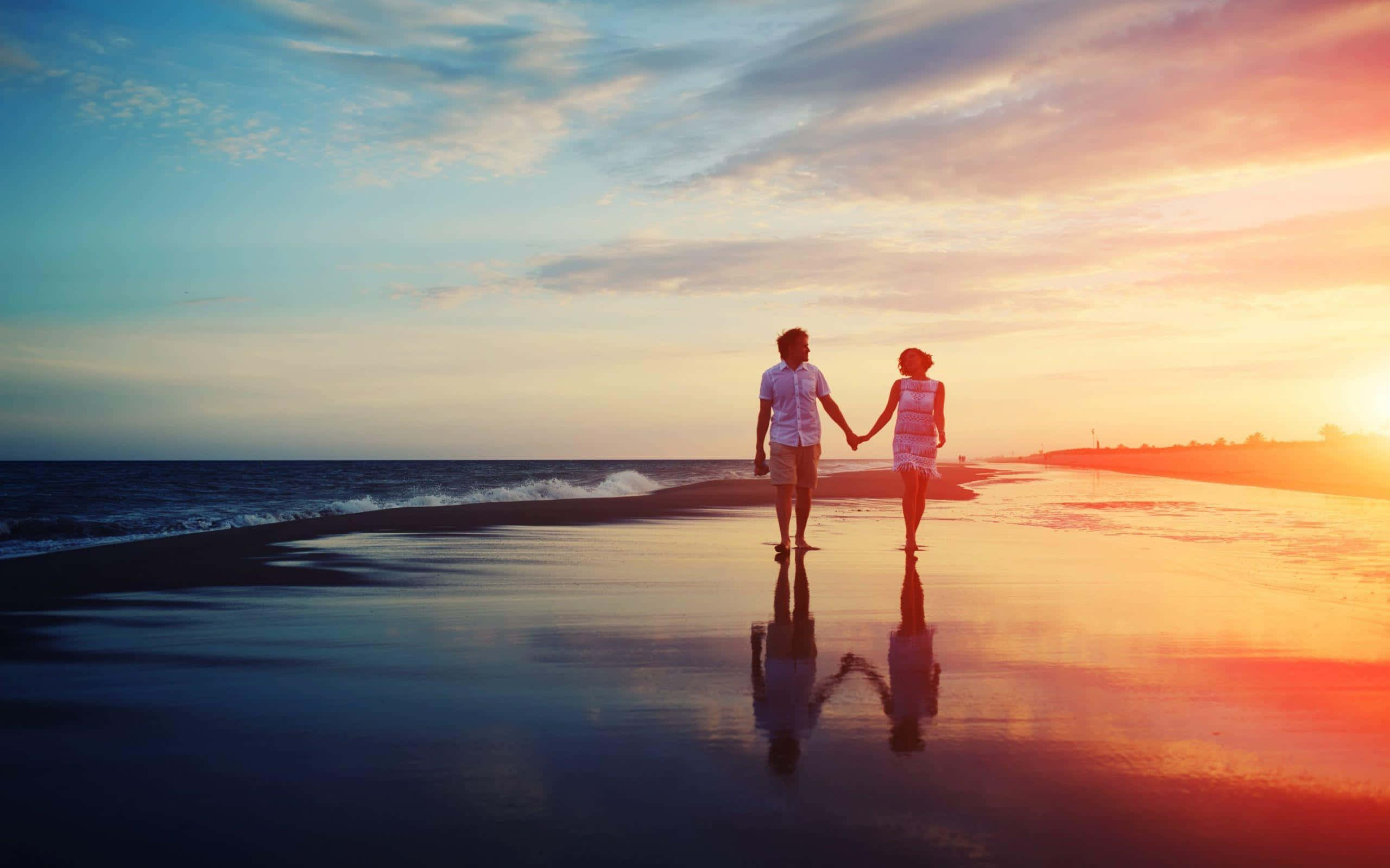 Beach Love 桌布 壁紙 — 一對情侶在日落時分手牽手沿著濕漉漉的海灘漫步，他們的倒影在淺水中閃爍。