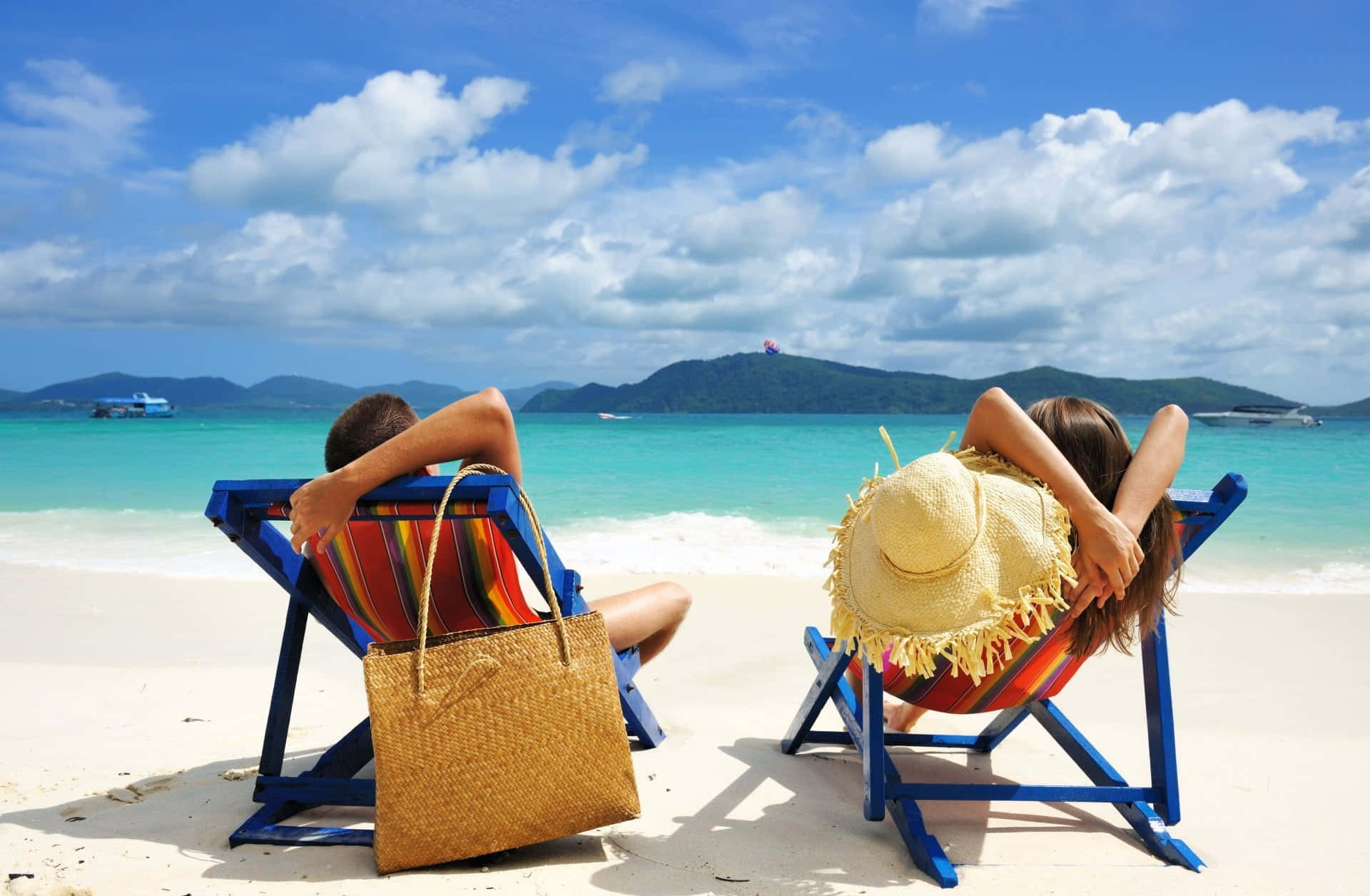Beach Bag 桌布 壁紙 — 一對情侶在白色沙灘上的彩色沙灘椅上放鬆，面對著碧綠的海水和遠處的綠色山丘，天空湛藍，雲朵蓬鬆。