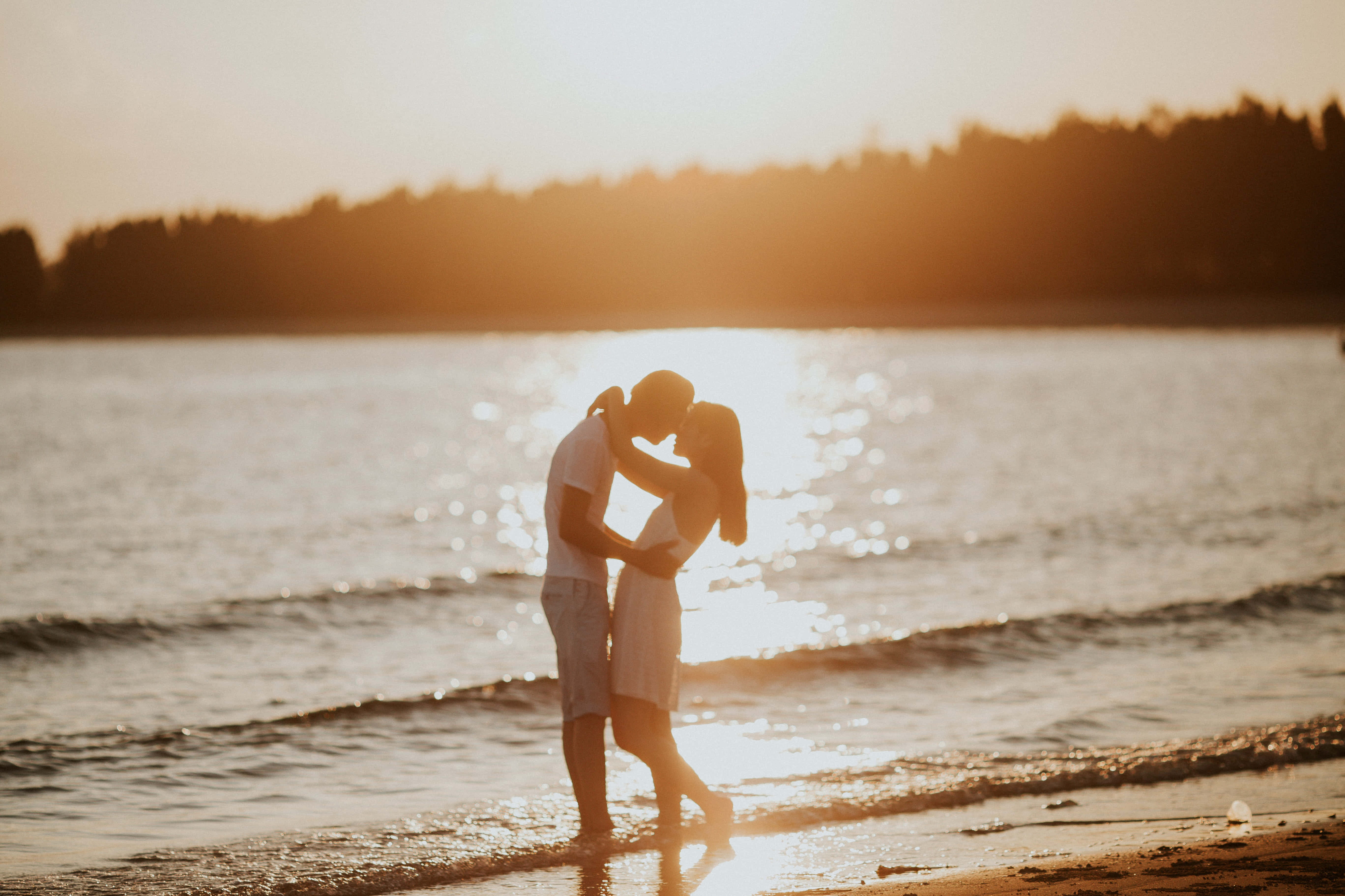 Beach Lovers 桌布 壁紙 — 一對情侶在陽光明媚的海灘上擁抱親吻，身後金色的夕陽在水面上閃爍。