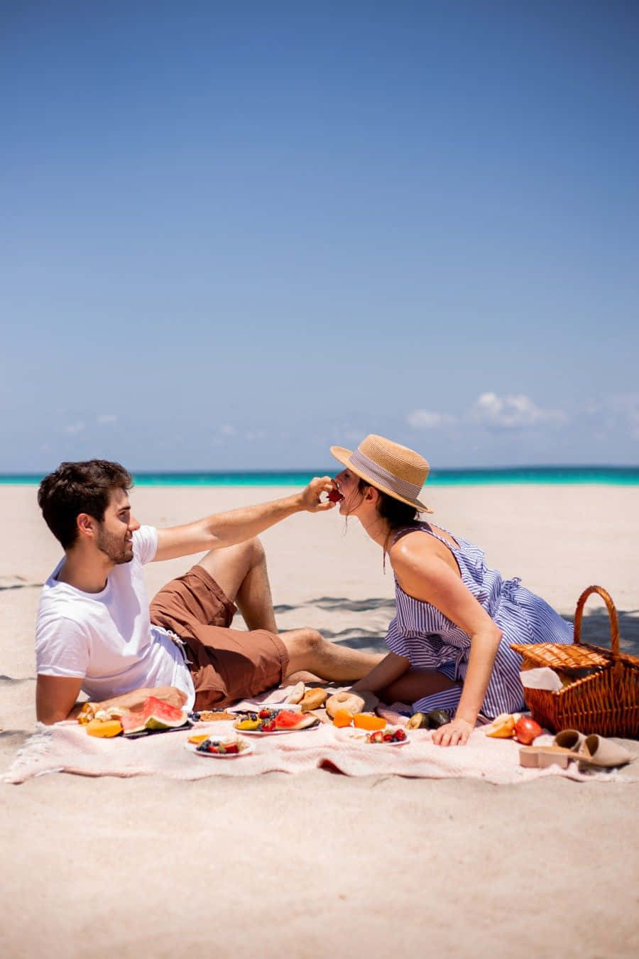 Beach Picnic 桌布 壁紙 — 一對情侶在陽光明媚的海灘野餐中互相餵食西瓜，背景是碧綠的海洋，分享著嬉戲的時刻。