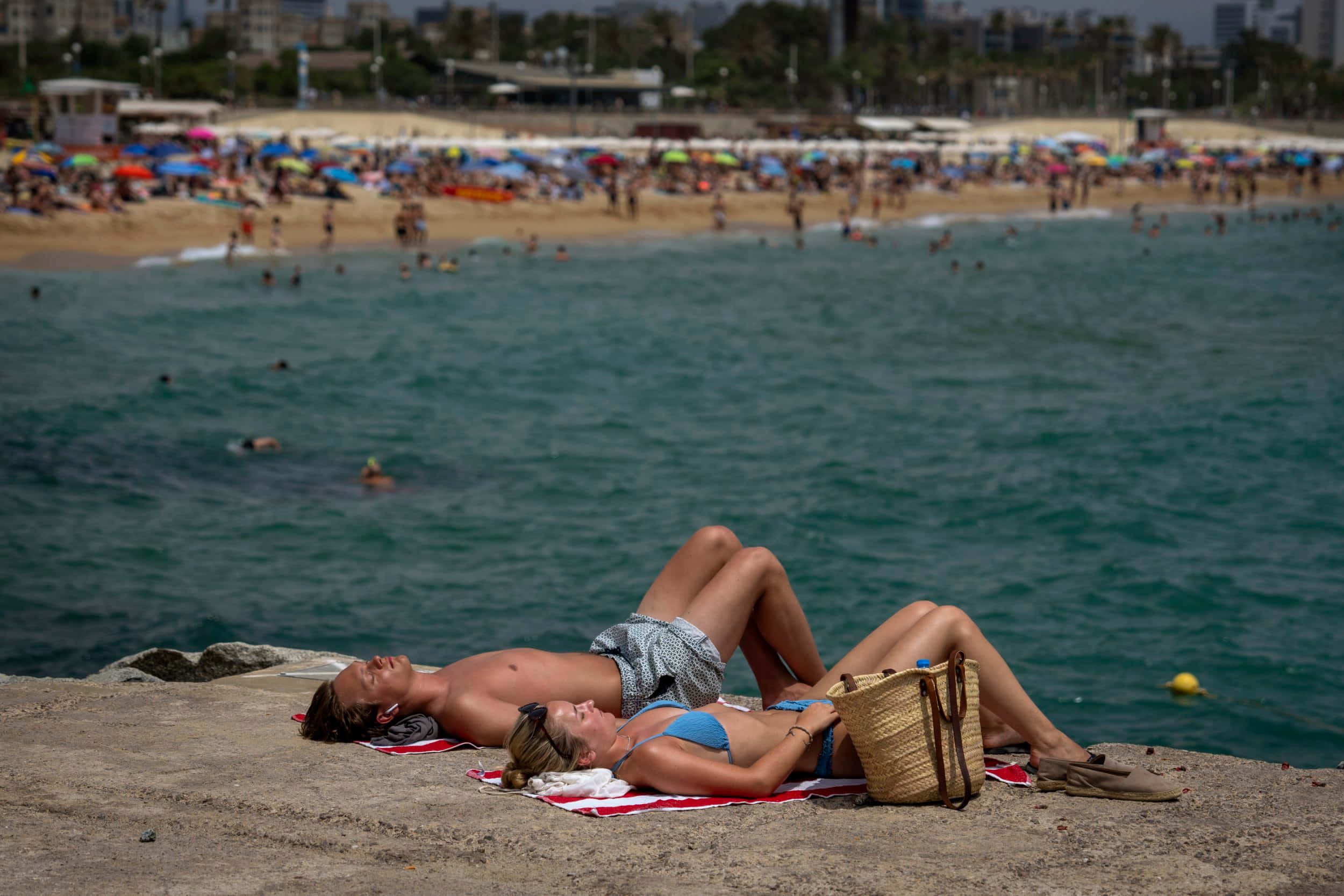 Spanish Beach 桌布 壁紙 — 一對情侶在陽光普照的懸崖上休息，俯瞰著繁忙的海灘，碧綠的海水和遠處的城市景觀。