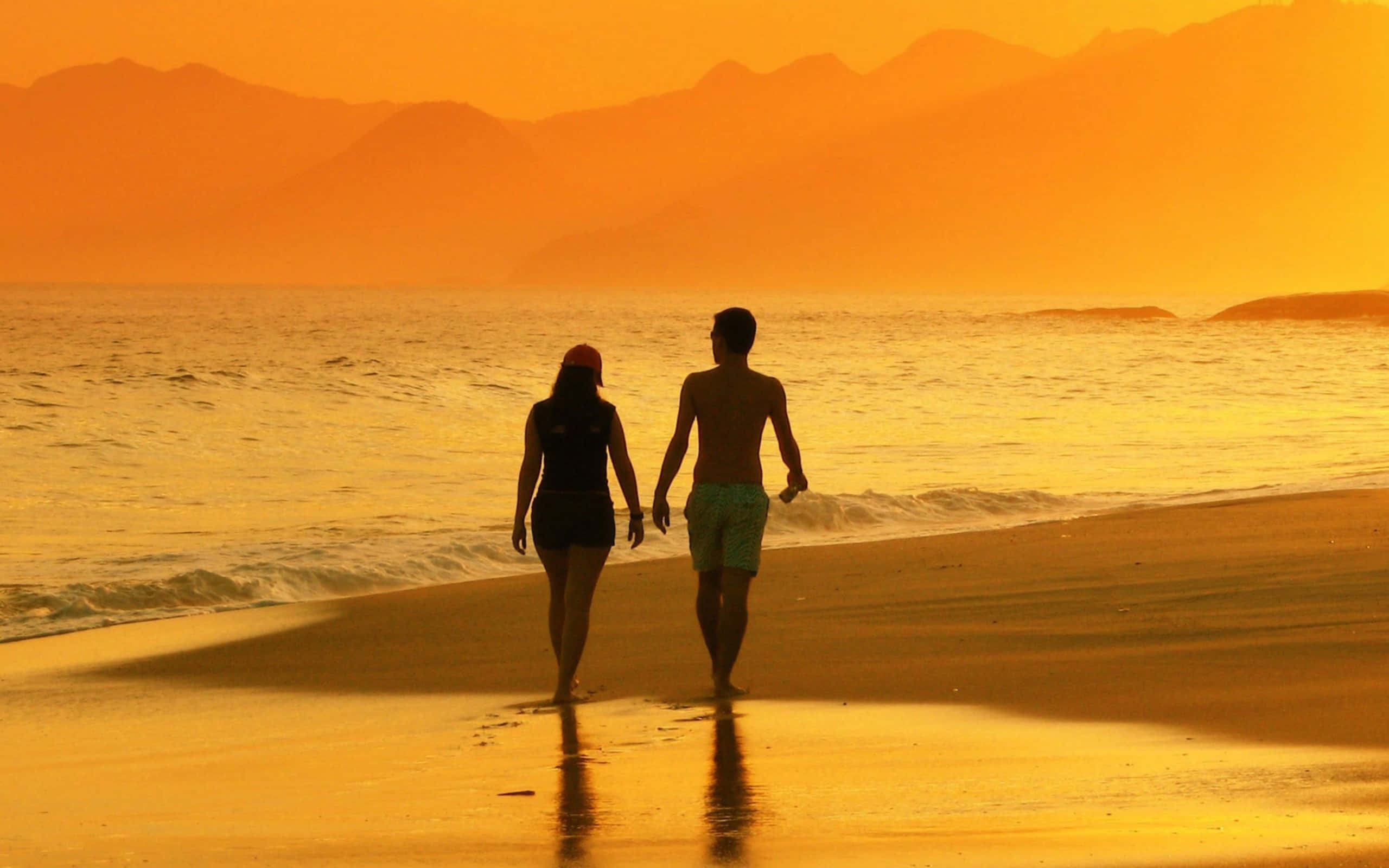 Beach Lovers 桌布 壁紙 — 一對情侶手拖手在日落時份沿著金色沙灘漫步，背景是剪影般的山巒。