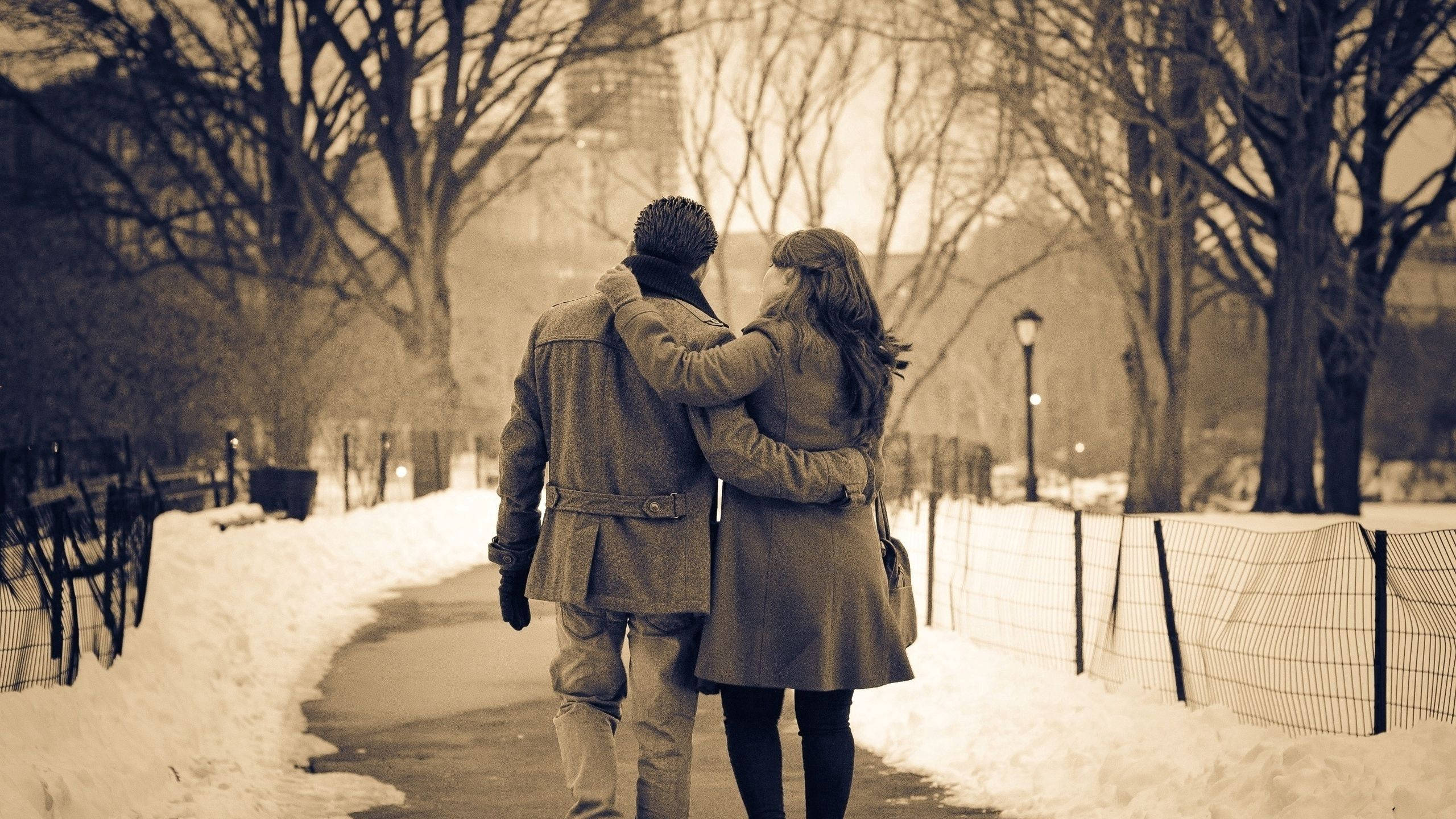 Girlfriend 桌布 壁紙 — 一對情侶手挽手走在白雪覆蓋的公園小徑上，兩旁是光禿禿的樹木，籠罩在柔和冬日陽光下。