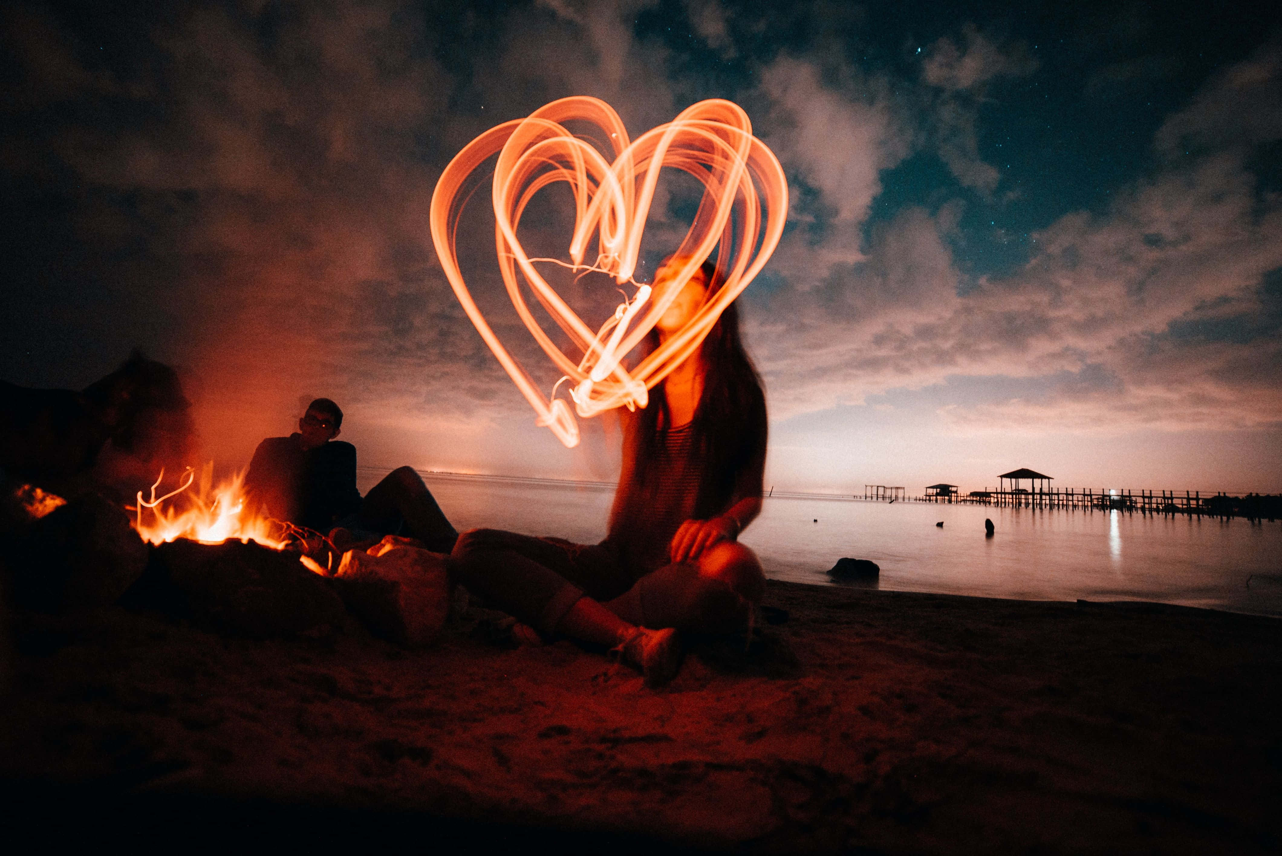 Beach Bonfire 桌布 壁紙 — 一對情侶晚上坐在海灘的營火旁，一名女子在星空下用光在他們上方畫出一個發光的愛心。
