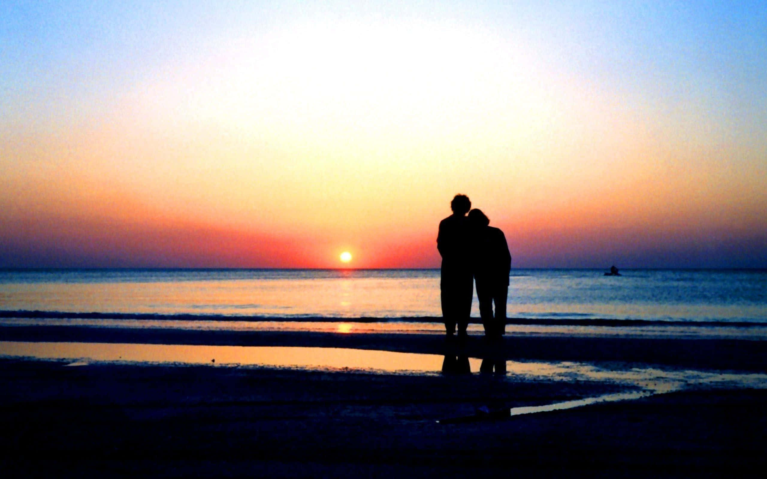 Beach Love 桌布 壁紙 — 一對情侶的剪影在海灘上擁抱，夕陽落在平靜的海面上，投射出溫暖的橙色和藍色色調。