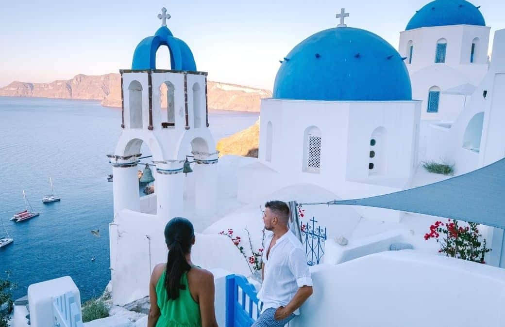 Greek Island 桌布 壁紙 — 一對情侶站在白色懸崖上，俯瞰愛琴海，藍頂教堂和帆船在晴朗的天空下點綴著地平線。