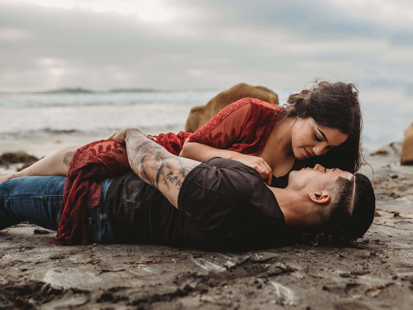 Beach Love 桌布 壁紙 — 一對情侶躺在濕漉漉的沙灘上，身後是海浪和陰天，分享親密時刻。
