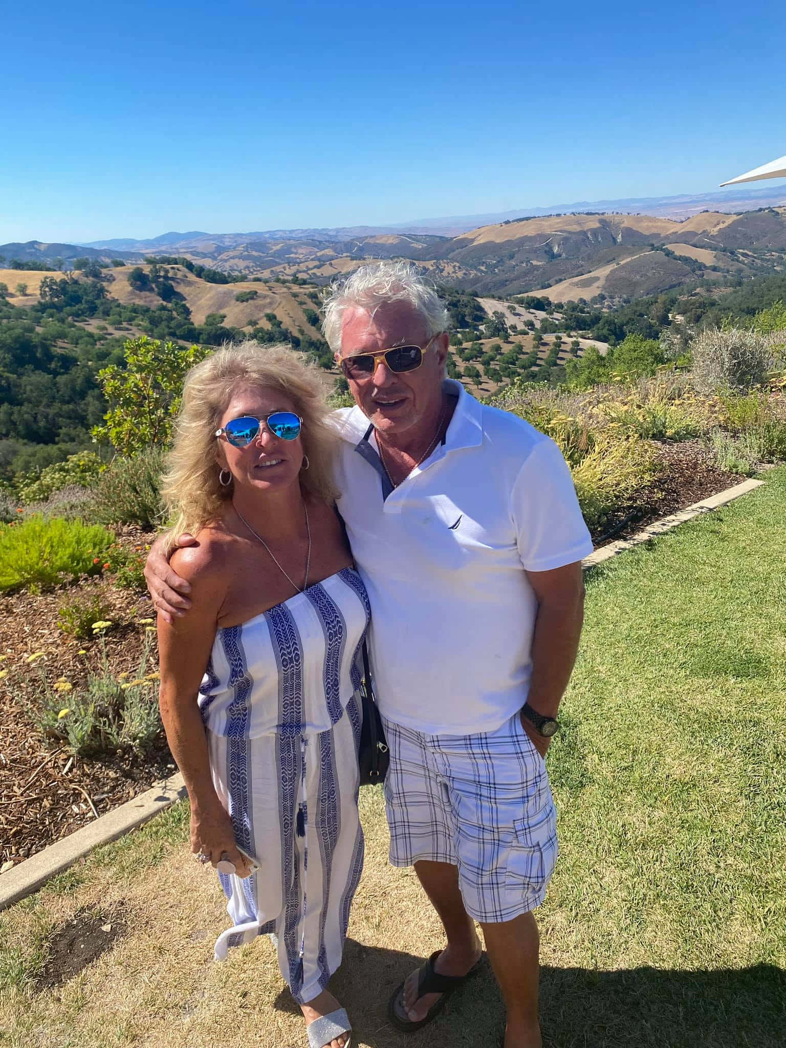 Tom Berenger 桌布 壁紙 — 一對戴著太陽眼鏡、笑容滿面的情侶，在陽光明媚的山坡上擺姿勢，俯瞰著連綿的金色山丘和遠處的山脈，天空清澈湛藍。