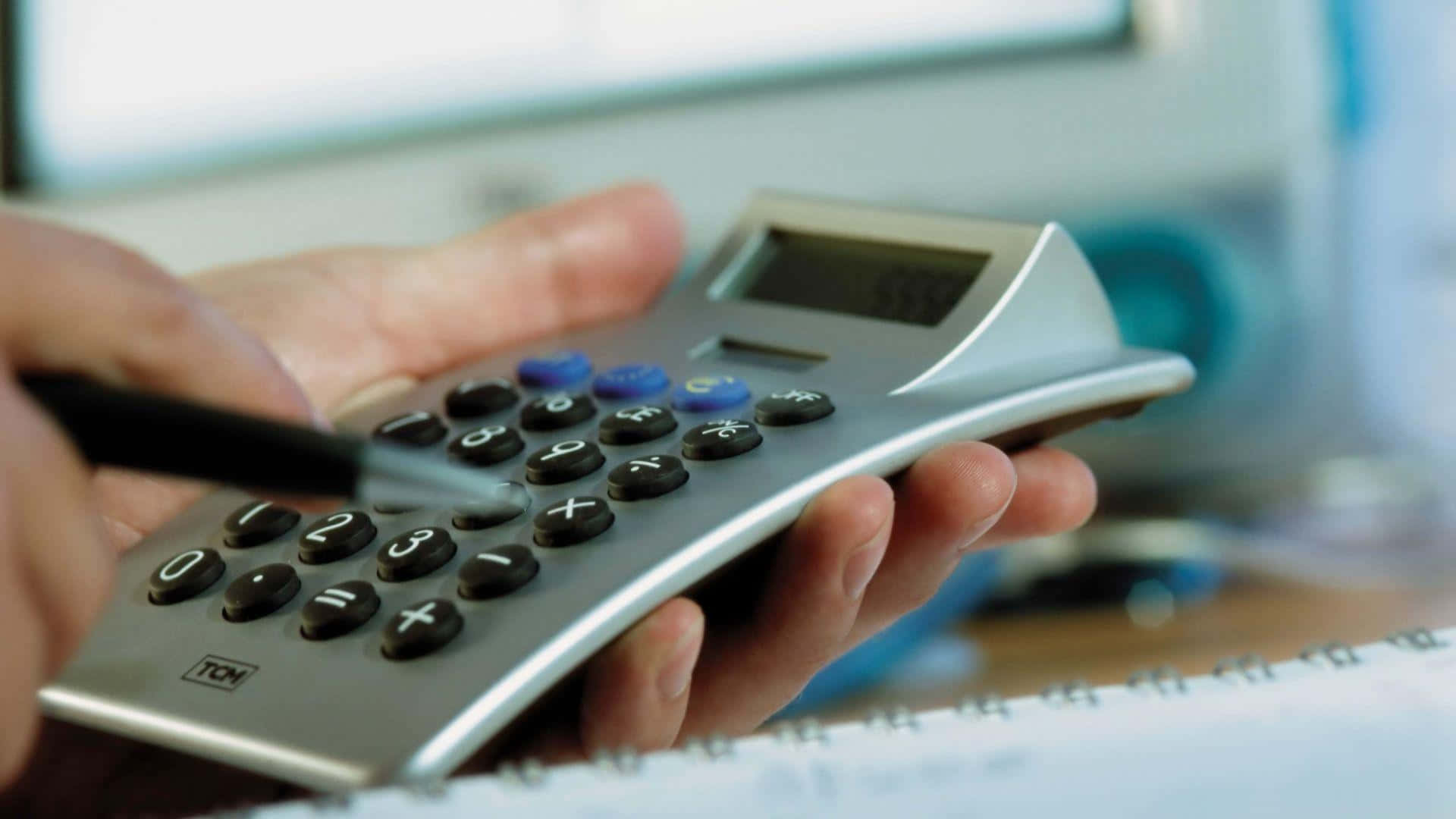 Calculator 桌布 壁紙 — 一對手持銀色計算機的手部特寫，筆尖懸浮在按鈕上方，暗示財務計算或預算規劃。