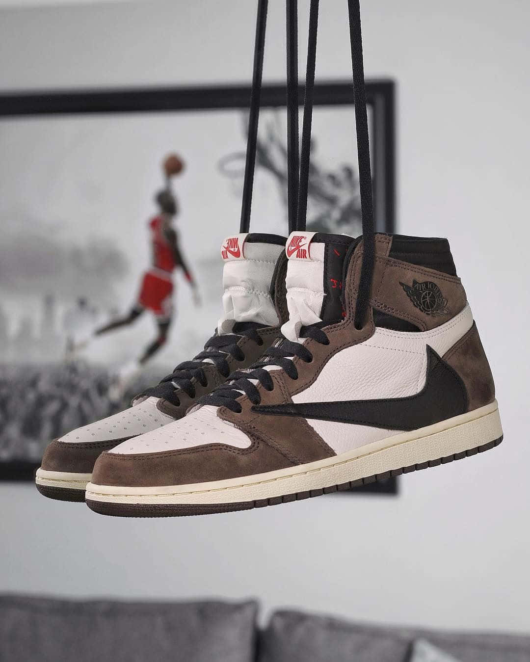 Travis Scott Jordan 1 桌布 壁紙 — 一對棕白色高筒運動鞋懸掛在鞋帶上，靠著一面牆，牆上掛著一幅裝裱的米高佐敦入樽照片。