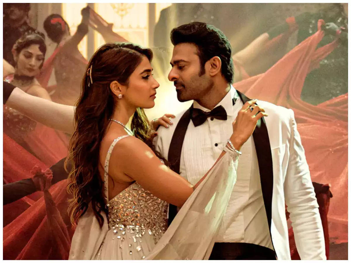 Prabhas 桌布 壁紙 — 一對穿著正式服裝的迷人夫婦，在金色燈光的舞台場景中，於充滿活力的紅色舞蹈團中分享親密時刻。