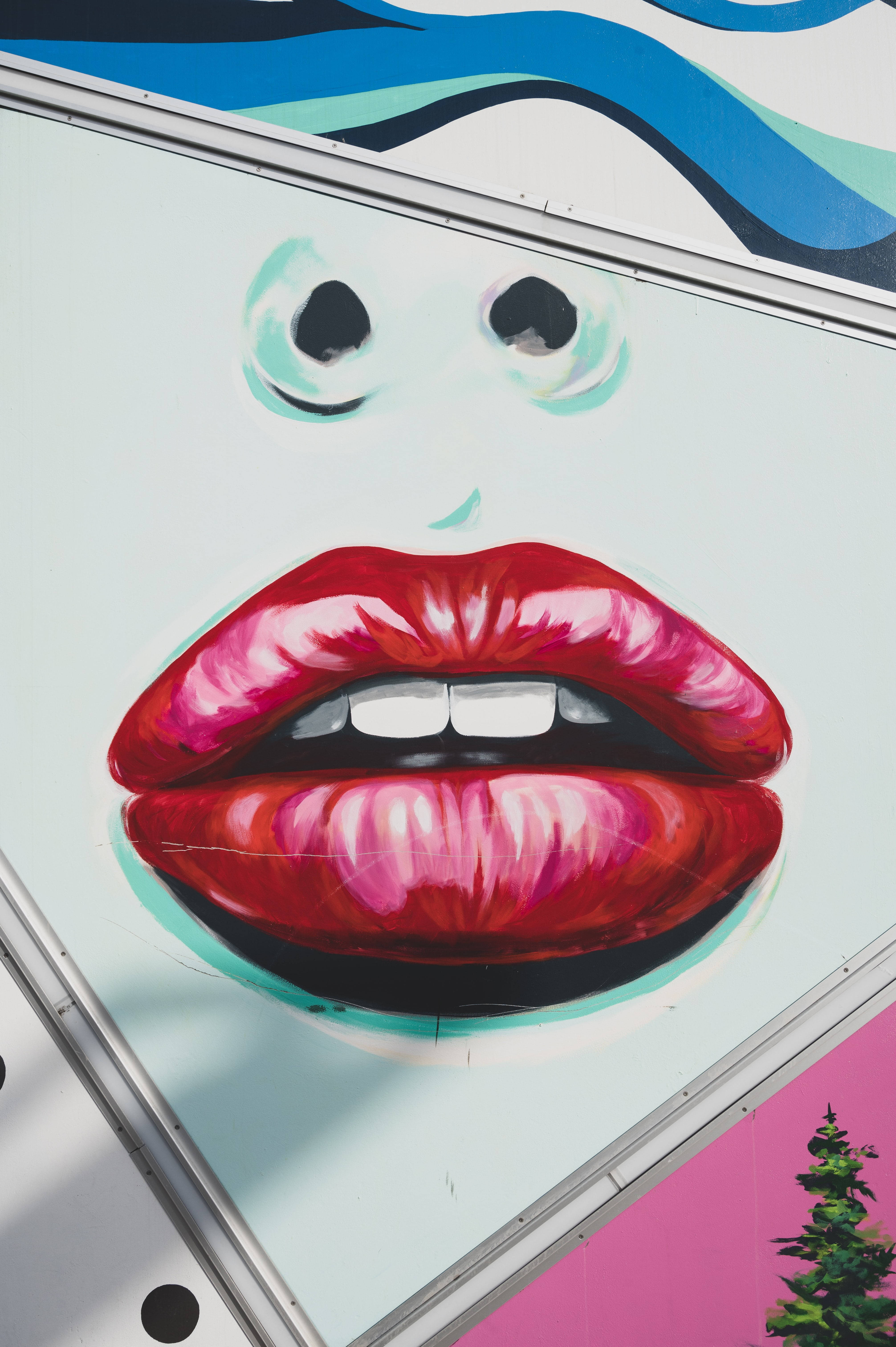 Art iPhone 桌布 壁紙 — 一幅充滿活力的塗鴉壁畫，描繪了一個風格化的面孔，帶有鮮豔的紅唇和大眼睛，畫在傾斜的牆壁上。