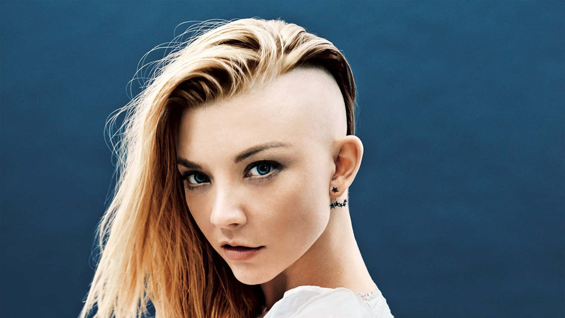 natalie dormer 桌布 壁紙 — 一幅引人注目的肖像，一位年輕女性擁有大膽的金色漸層髮型和銳利的藍色眼睛，背景為深藍色。