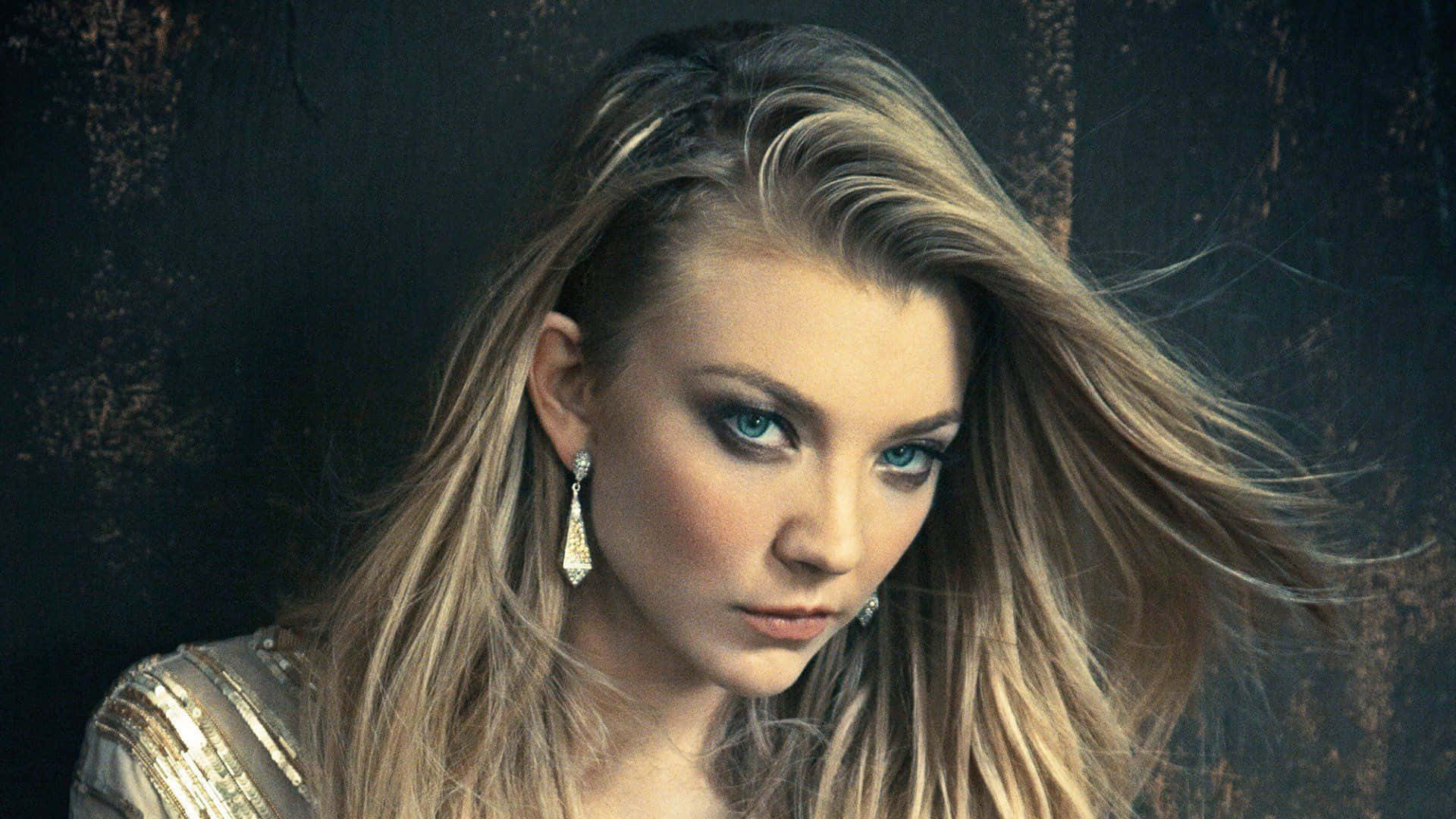 natalie dormer 桌布 壁紙 — 一幅引人注目的肖像，一位擁有飄逸金髮和銳利藍眼睛的女性，背景是深色紋理牆壁。