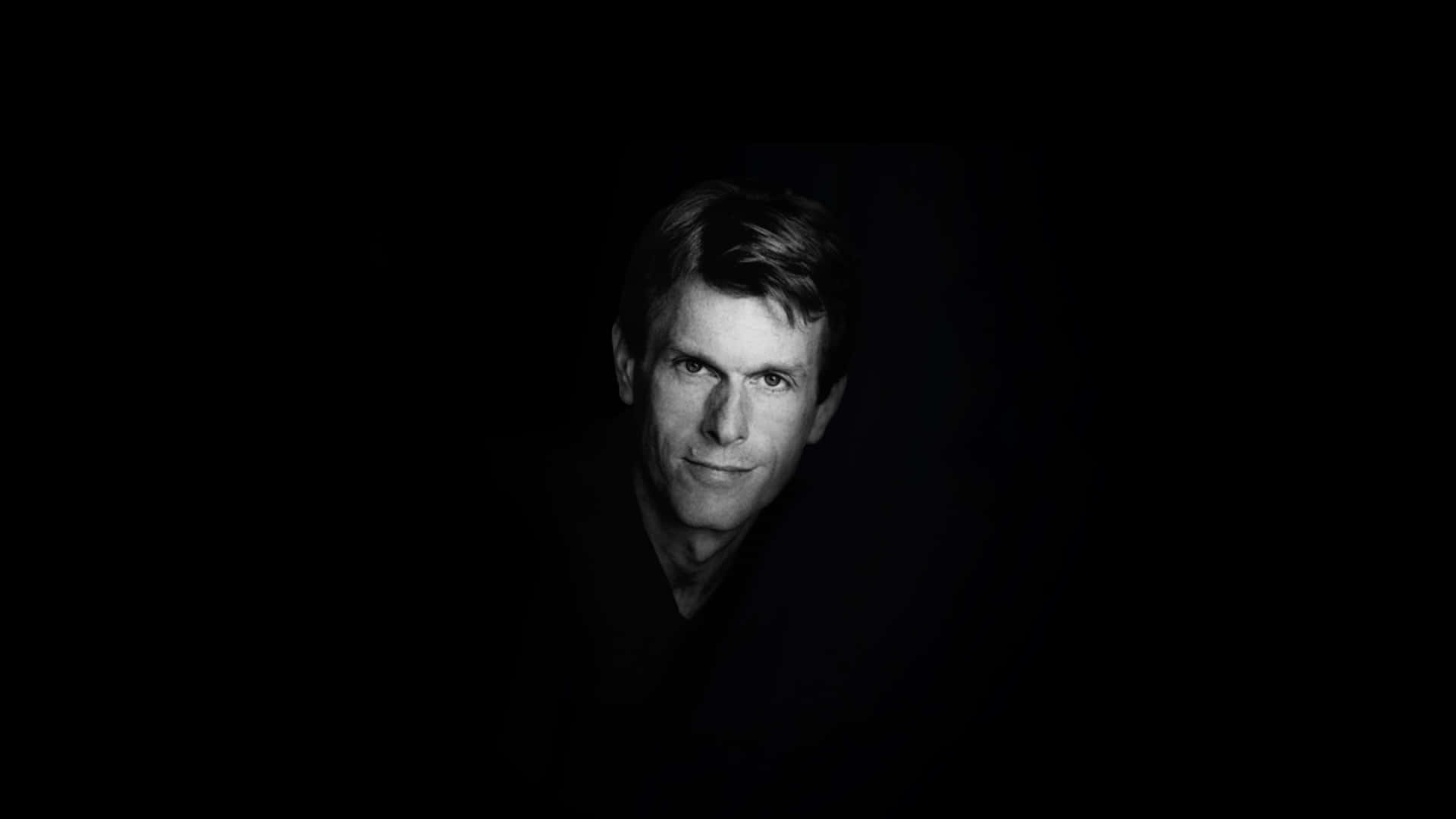 Kevin Conroy 桌布 壁紙 — 一幅強烈黑白肖像，捕捉一位眼神銳利、帶着微妙微笑的男子，背景完全漆黑。