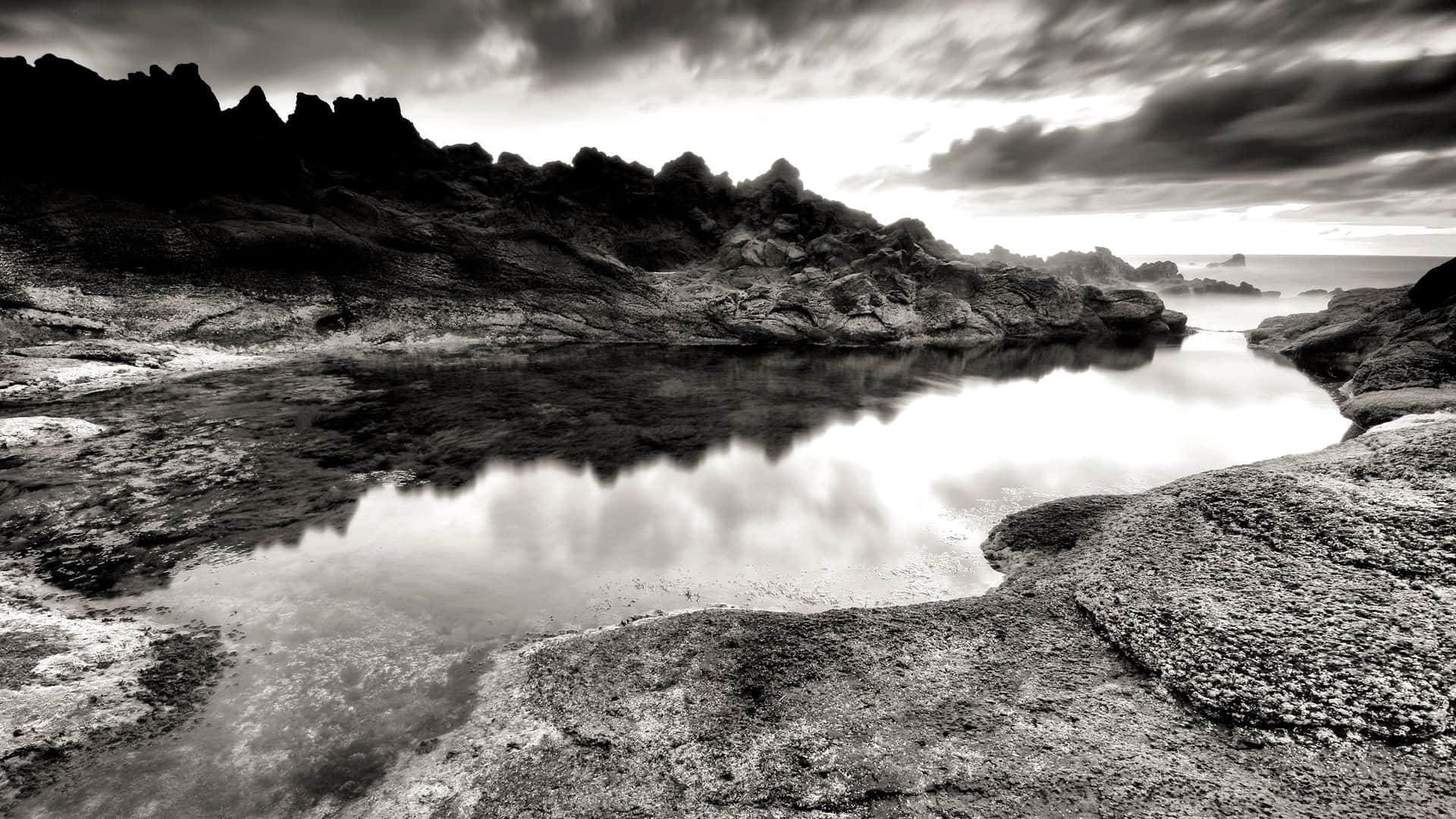 Black And White Nature 桌布 壁紙 — 一幅戲劇性的黑白風景，顯示在多雲的天空下，參差不齊的岩石環繞著平靜的倒影池。