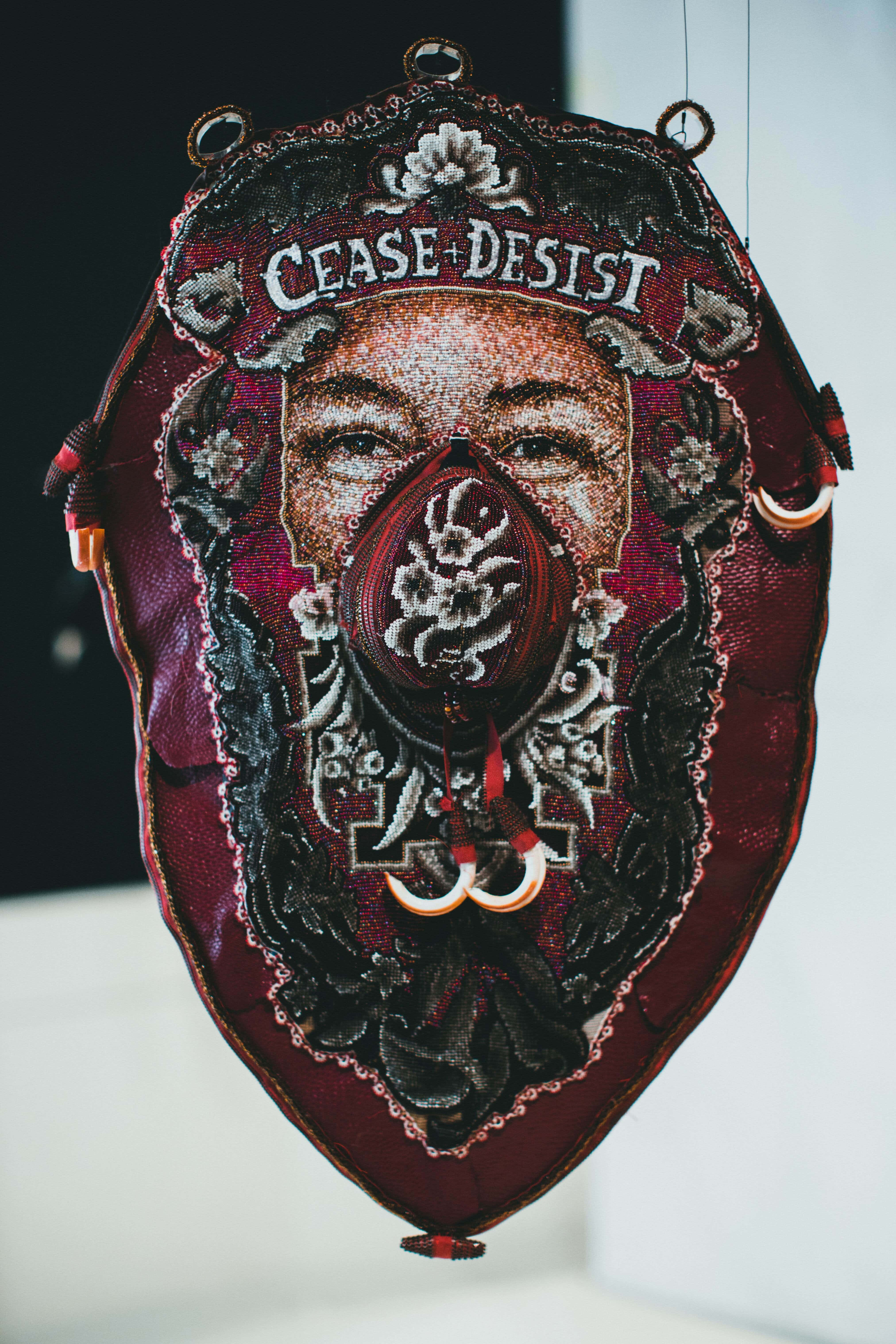 shield 桌布 壁紙 — 一幅掛起的刺繡紡織藝術品，描繪了一張帶有「CEASE DESIST」文字和深紅色及黑色花卉圖案的臉孔。