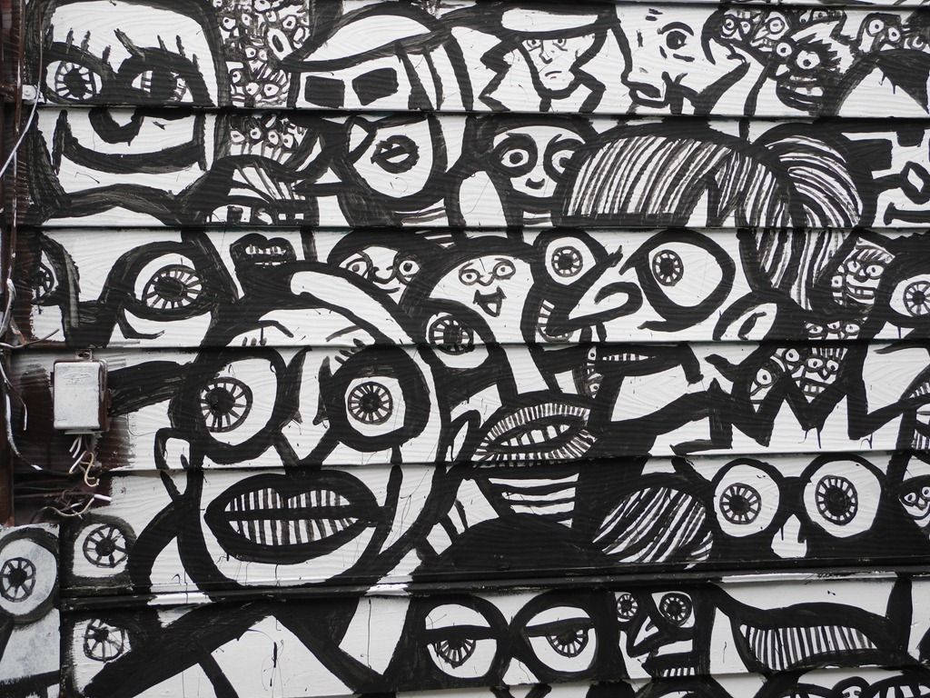 Black And White Graffiti 桌布 壁紙 — 一幅混亂的黑白壁畫，描繪了風格化的卡通面孔，帶有誇張的眼睛和嘴巴，覆蓋在風化的木質牆板上。