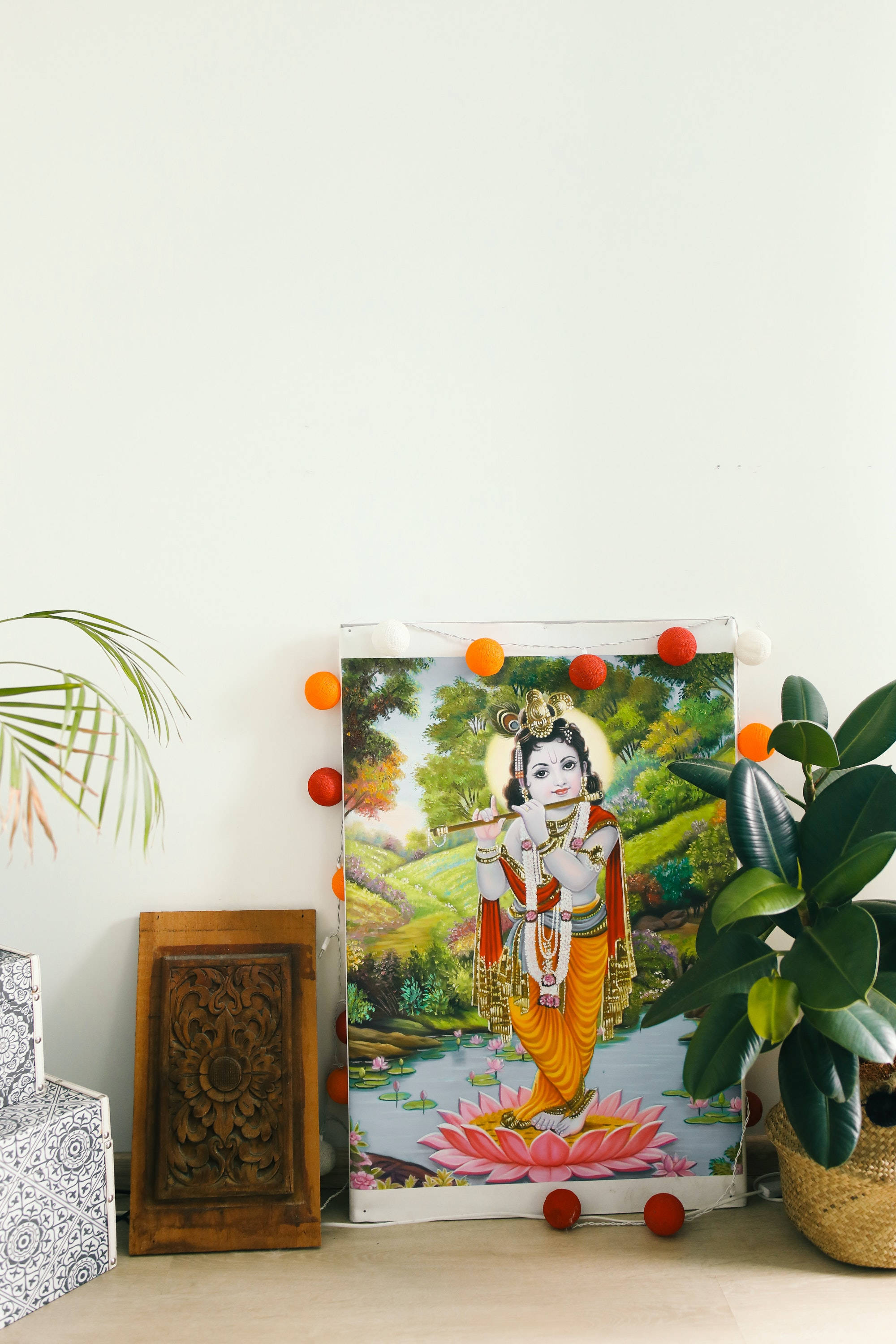 Lord Shiva 桌布 壁紙 — 一幅色彩鮮豔的畫作，描繪克里希納主在蓮花上吹笛，裱框並用橙色絨球裝飾，掛在白色牆上，旁邊有綠色植物。