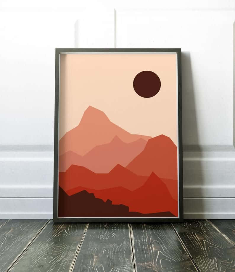 Minimalist Painting 桌布 壁紙 — 一幅裱框的極簡主義山脈藝術印刷品，帶有層疊的紅橙色山峰和淡蜜桃色天空中的暗色太陽，靠在白色牆壁上，下方是深色木地板。