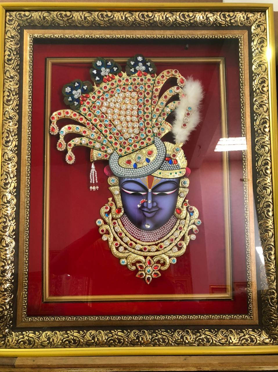 Shrinathji 桌布 壁紙 — 一幅鑲滿寶石的藍色神祇肖像，頭戴金色皇冠和羽毛，背景為深紅色，置於華麗的金色畫框中。