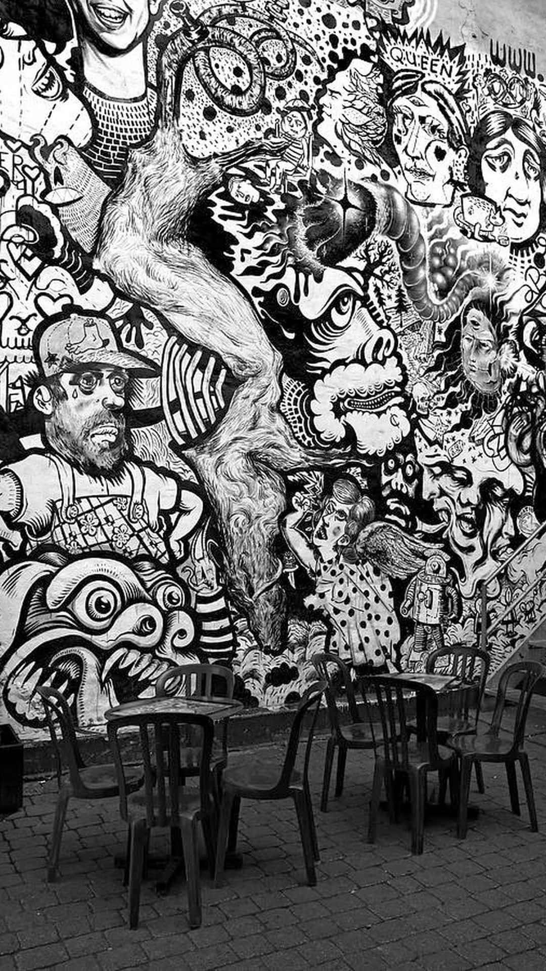 Black And White Graffiti 桌布 壁紙 — 一幅黑白壁畫，描繪混亂的卡通面孔和人物，覆蓋在戶外塑膠桌椅後的牆上。