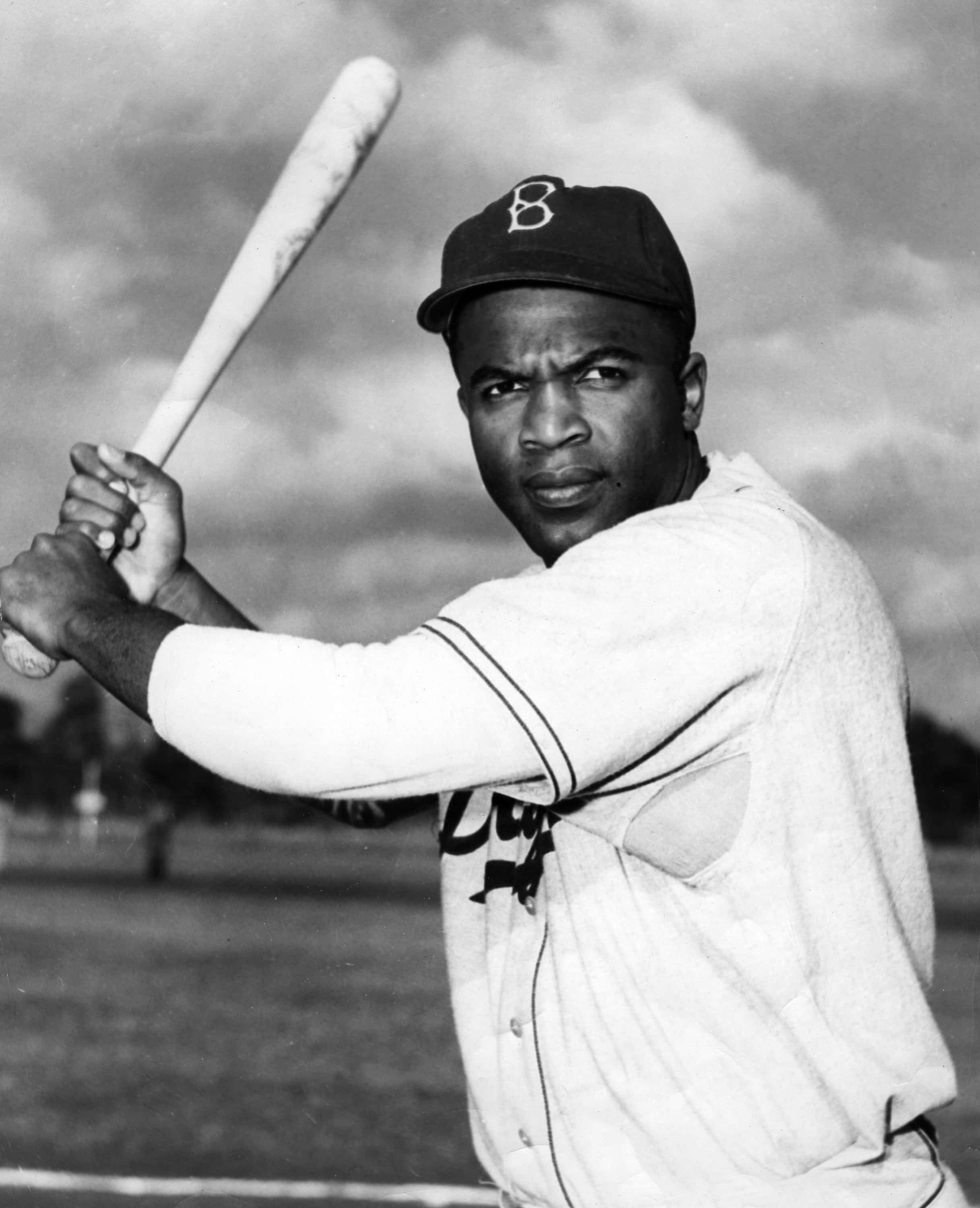 Jackie Robinson 桌布 壁紙 — 一幅黑白肖像，描繪一位身穿布魯克林隊服的堅定棒球運動員，在陰雲密布的天空下揮棒中途握著球棒。