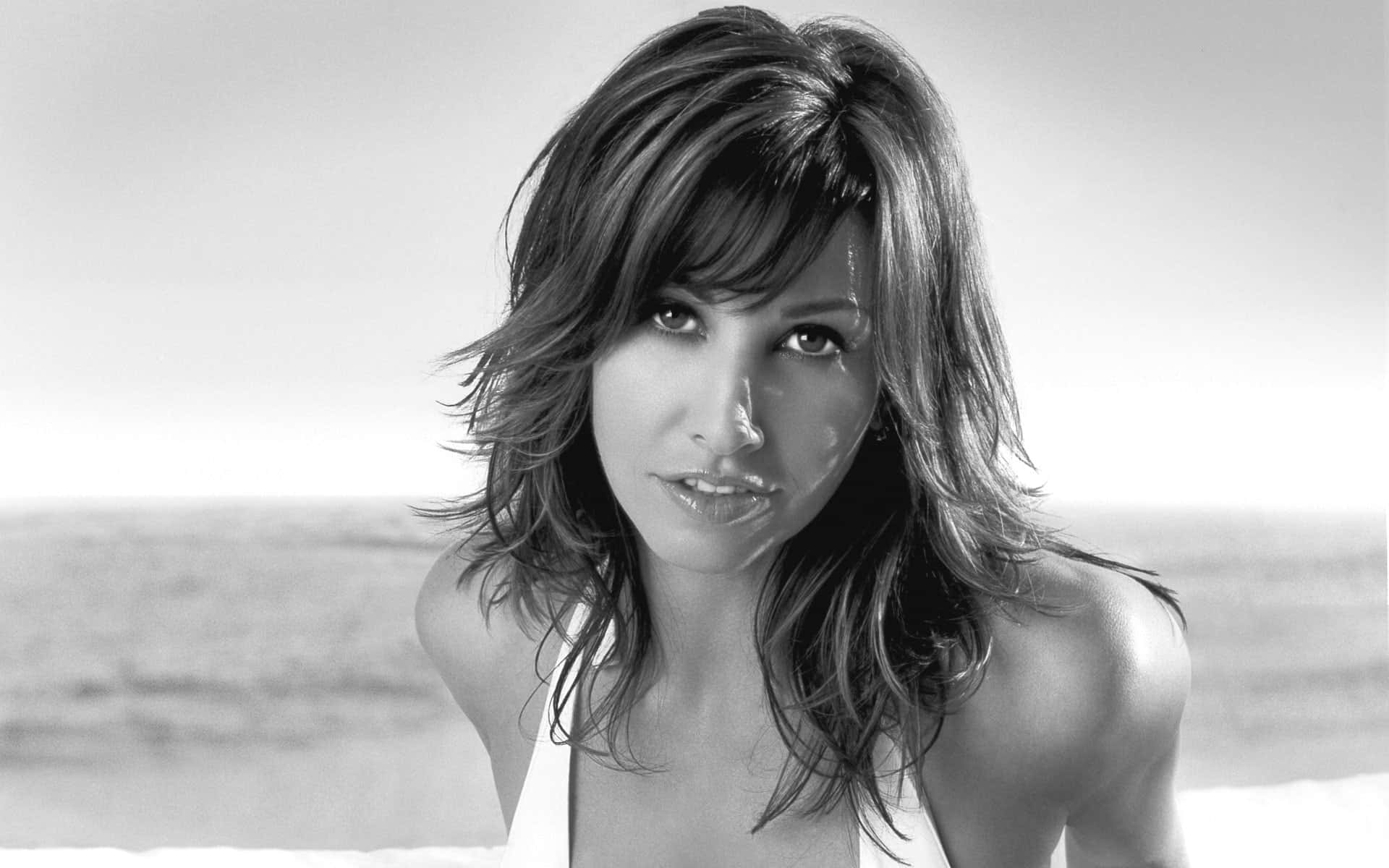 Gina Gershon 桌布 壁紙 — 一幅黑白肖像照，一名頭髮被風吹亂的女子在模糊的海洋背景下直視鏡頭。