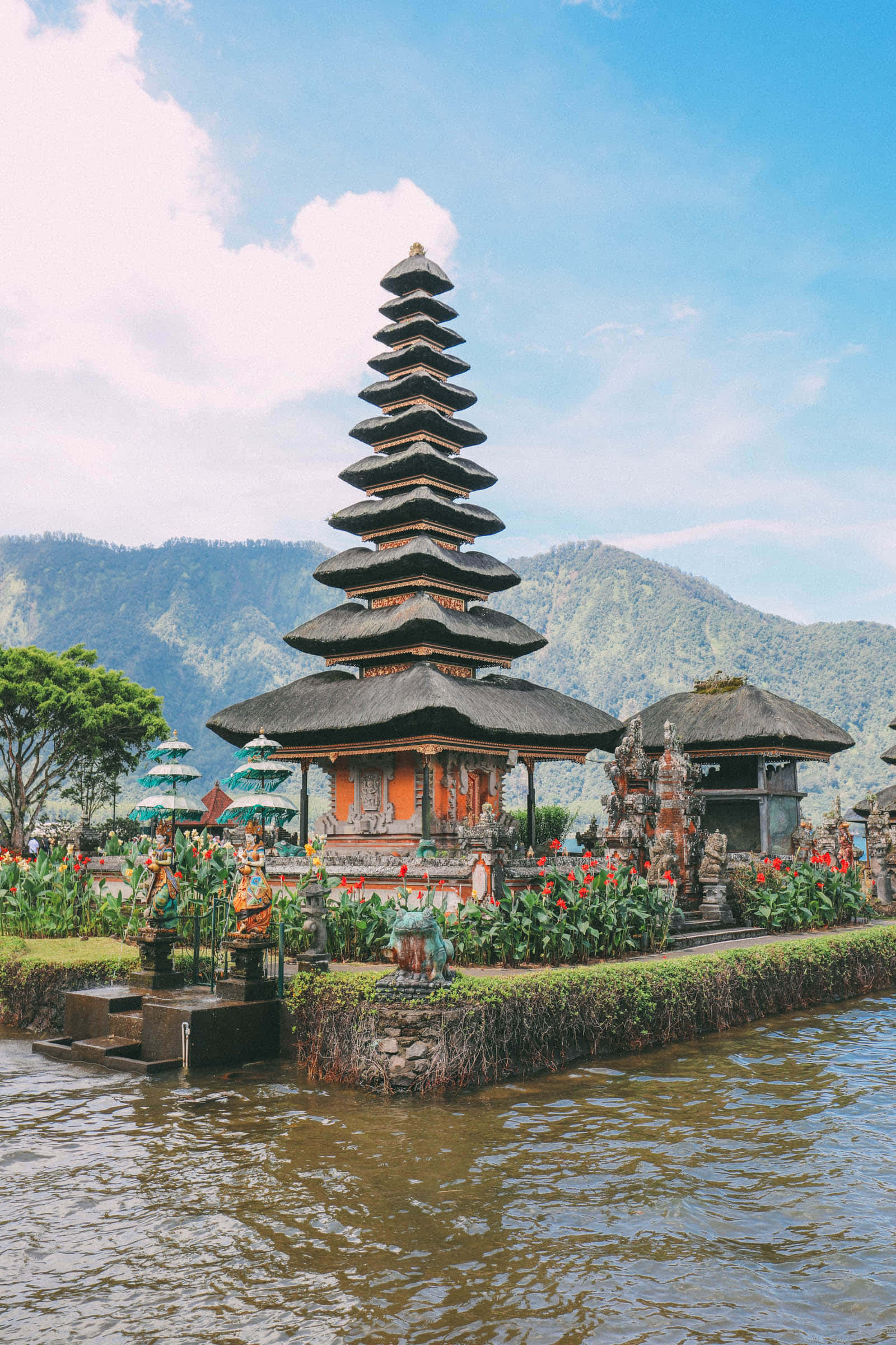 Bali 桌布 壁紙 — 一座傳統峇里島寺廟，擁有層疊屋頂，優雅地矗立於注滿水的蓮花池中，四周環繞著茂密的綠植和遠處的山巒，上方是明亮的藍天。