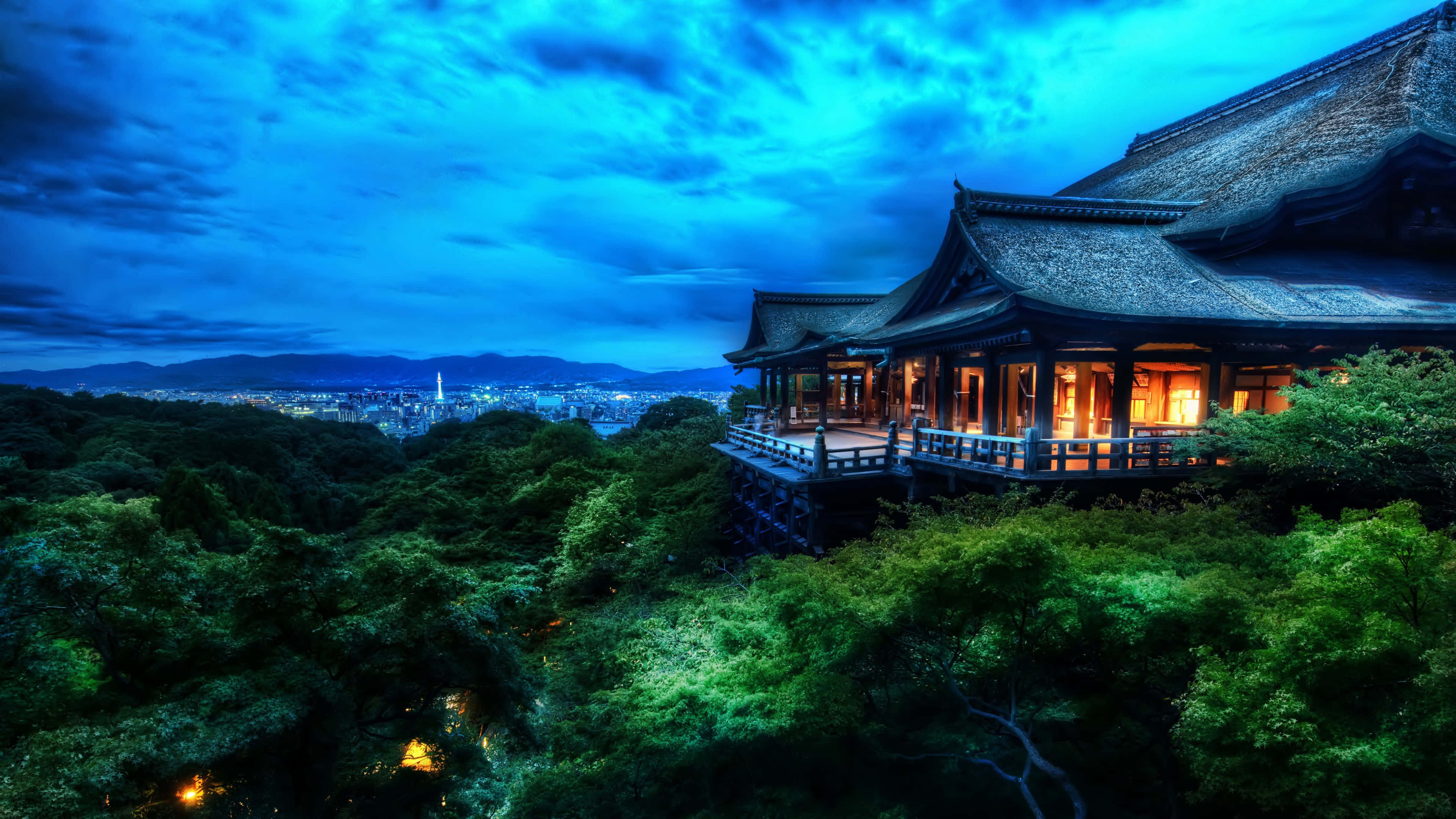 4K Ultra HD 桌布 壁紙 — 一座傳統日本寺廟坐落在林木茂密的山丘上，在深藍色的暮色天空和遠處的城市燈光映襯下，散發著溫暖的光芒。