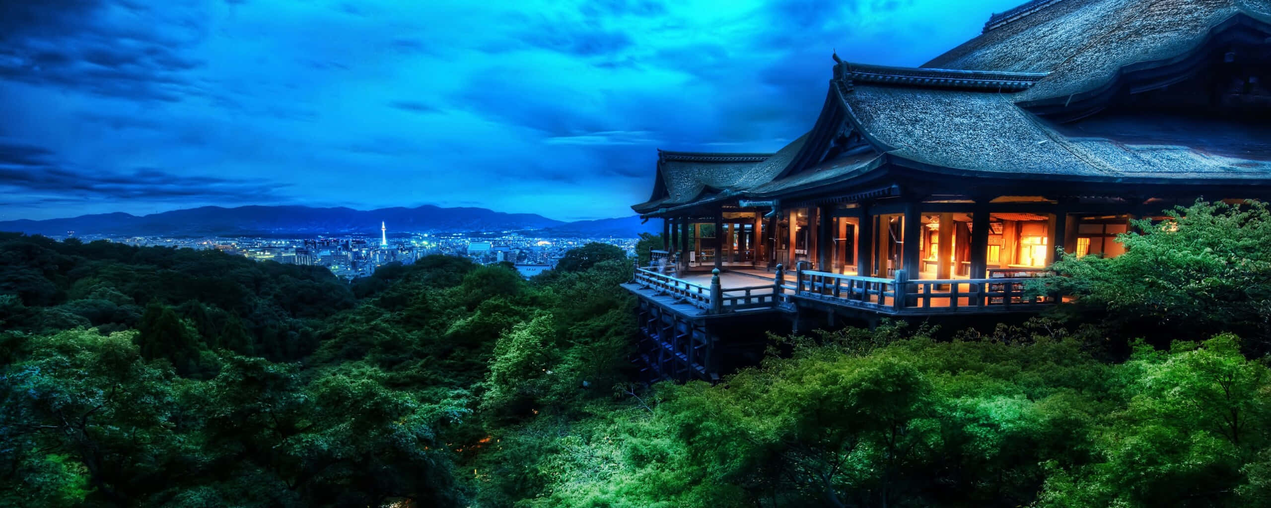 Feudal Japan 桌布 壁紙 — 一座傳統日本寺廟坐落在茂密的山坡上，在深藍色的暮色天空中溫暖地發光，俯瞰著遠處的城市景觀。