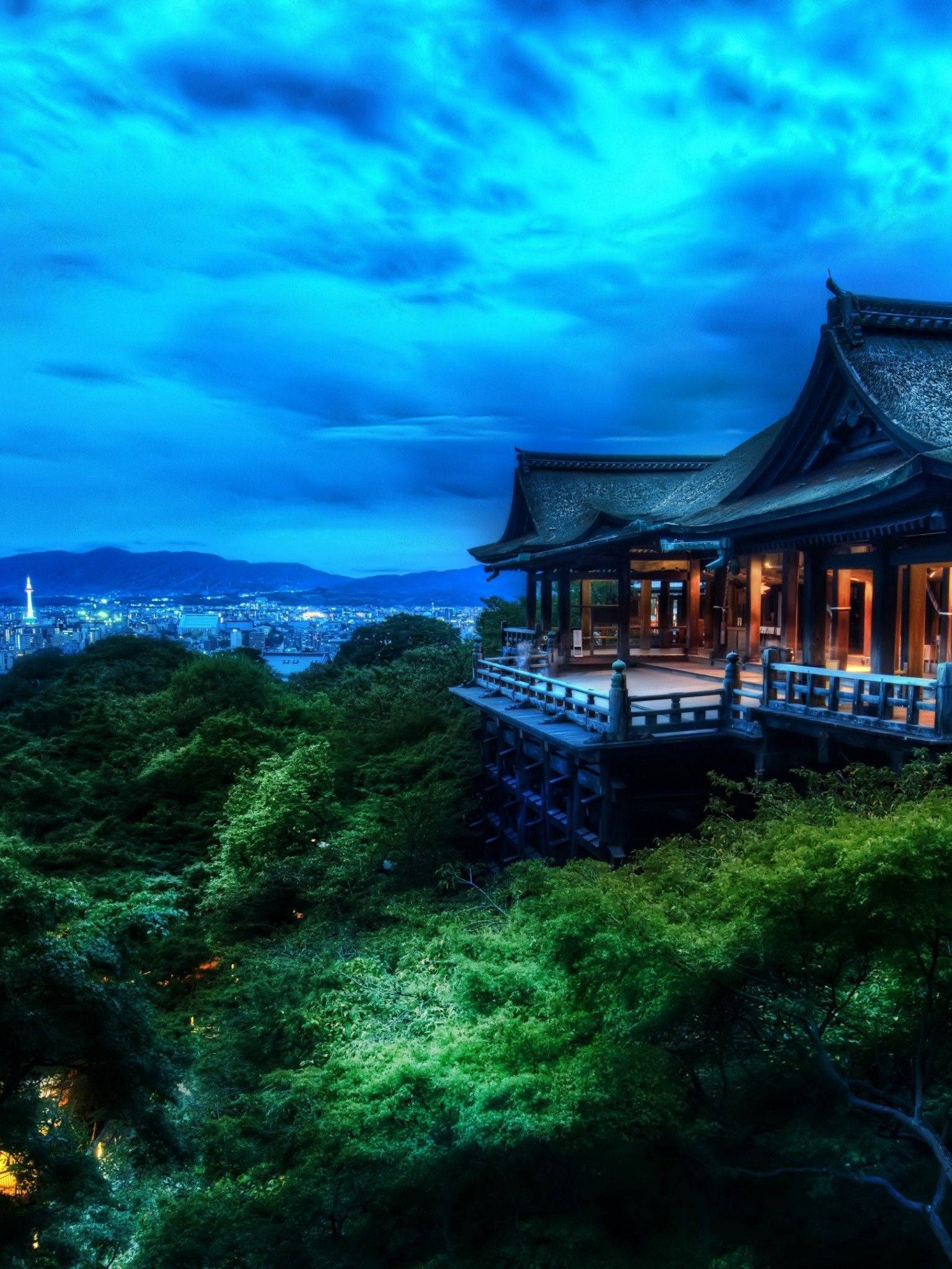 背景 桌布 壁紙 — 一座傳統木造寺廟坐落在茂密的綠樹之上，俯瞰著在戲劇性暮色天空下延伸的城市景觀。
