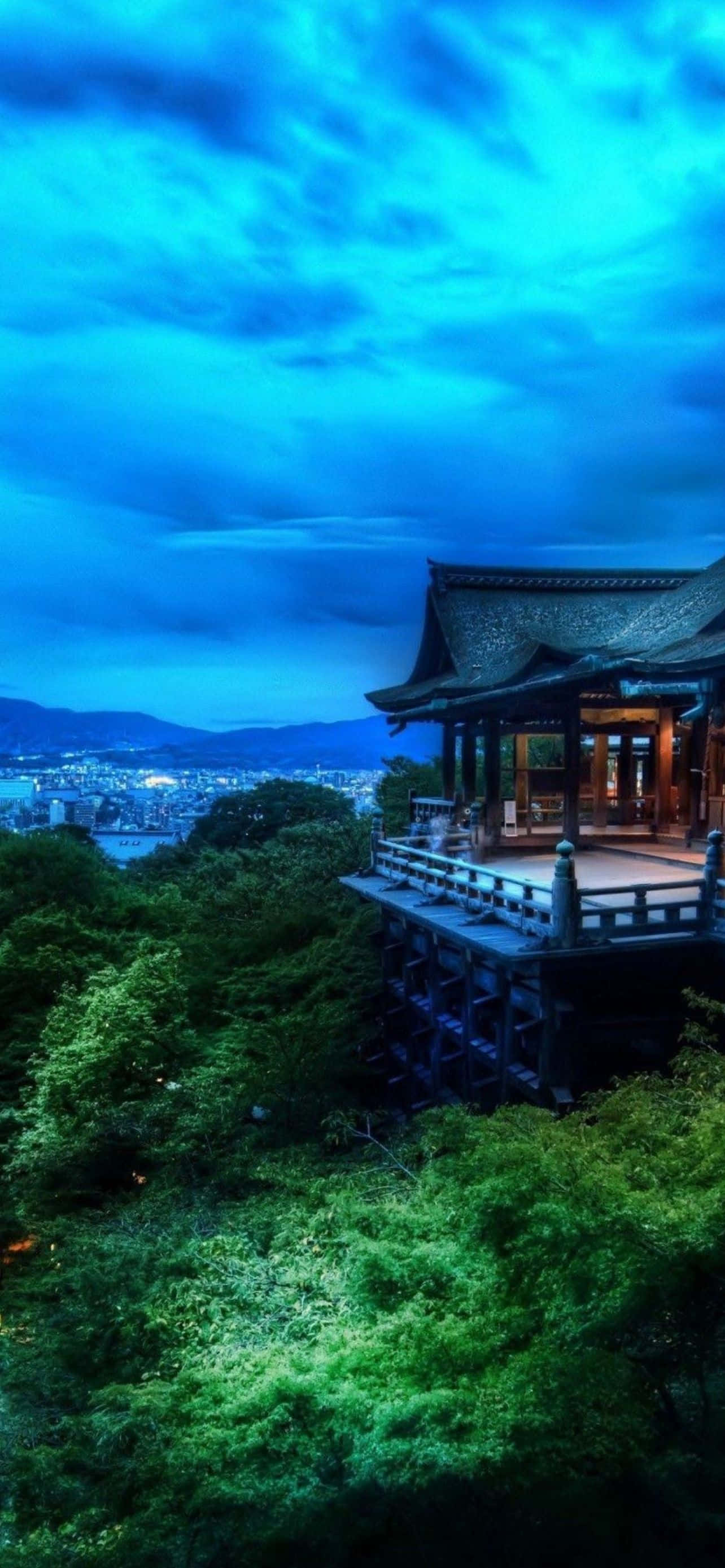 型格 iPhone 壁紙 桌布 壁紙 — 一座傳統木造寺廟坐落在蒼翠的山丘上，俯瞰著在戲劇性暮色天空下延伸的城市。