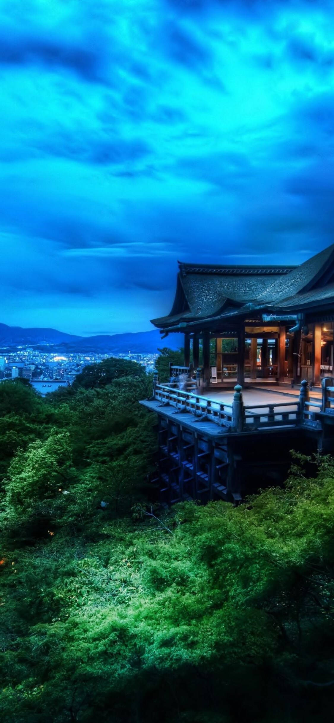 自然 桌布 壁紙 — 一座傳統木造寺廟坐落在鬱蔥的綠色山丘上，俯瞰著在戲劇性暮色天空下延伸的城市。