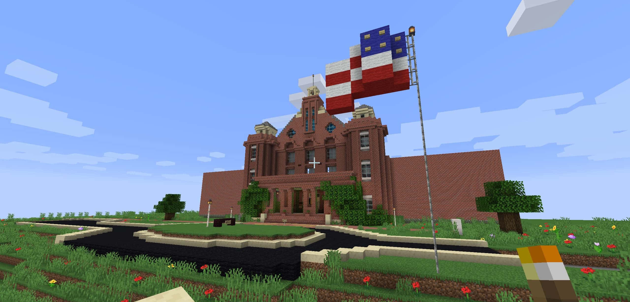Minecraft Building 桌布 壁紙 — 一座像素化的紅磚建築，飄揚著美國國旗，矗立在晴朗藍天下的草地上。