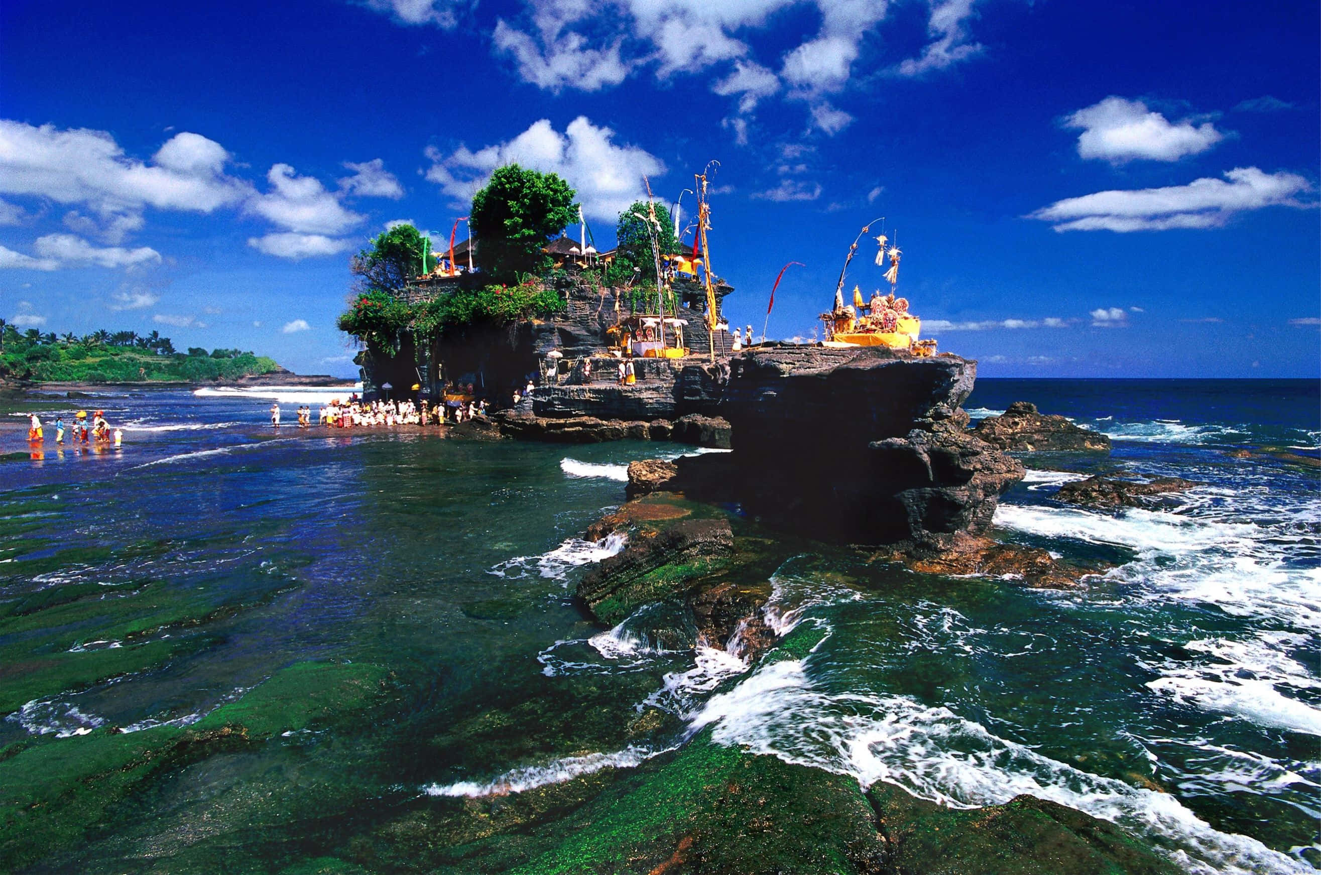 Bali Beach 桌布 壁紙 — 一座充滿活力的寺廟坐落在岩石島上，周圍是碧綠的海水，上方是明亮的藍天，點綴著幾朵雲。