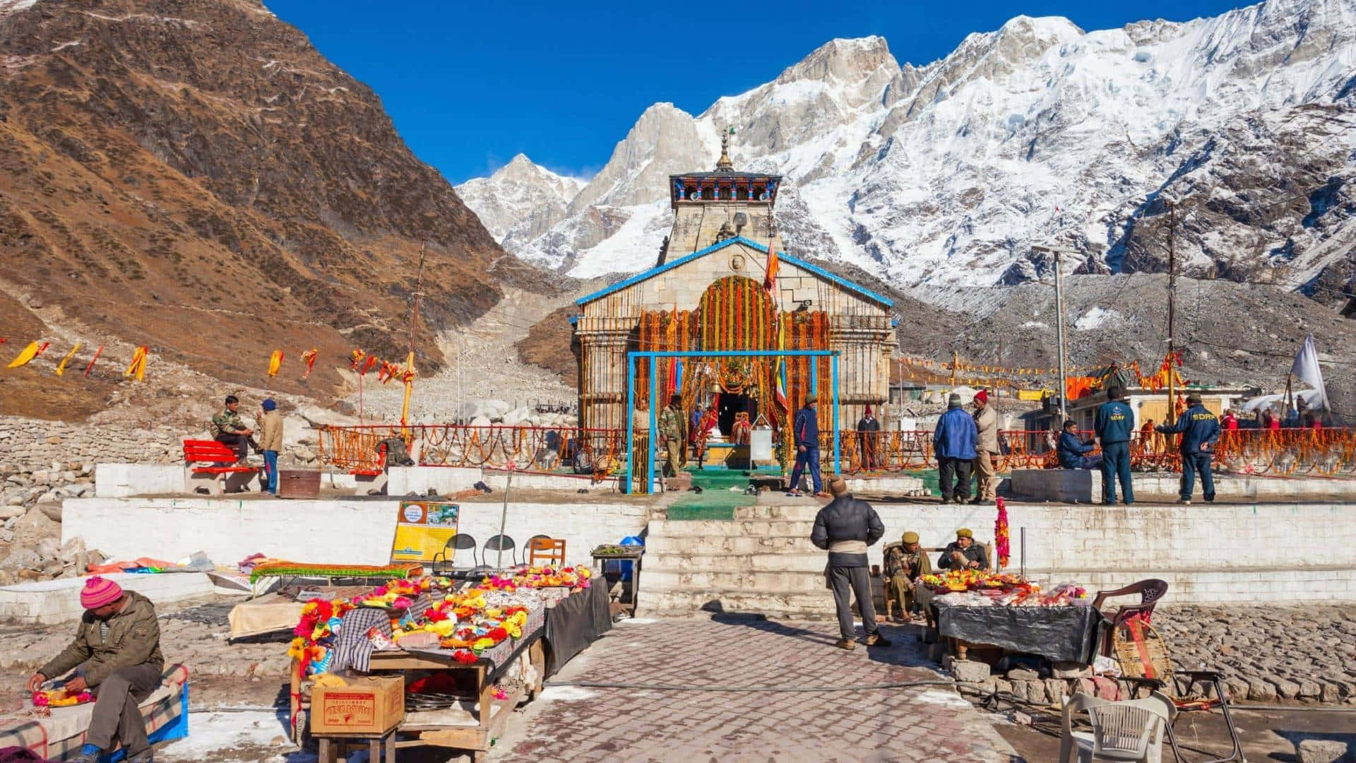 Kedarnath 桌布 壁紙 — 一座充滿活力的山間神社，裝飾著色彩繽紛的經幡和供品，矗立在晴朗藍天下高聳的雪峰前。