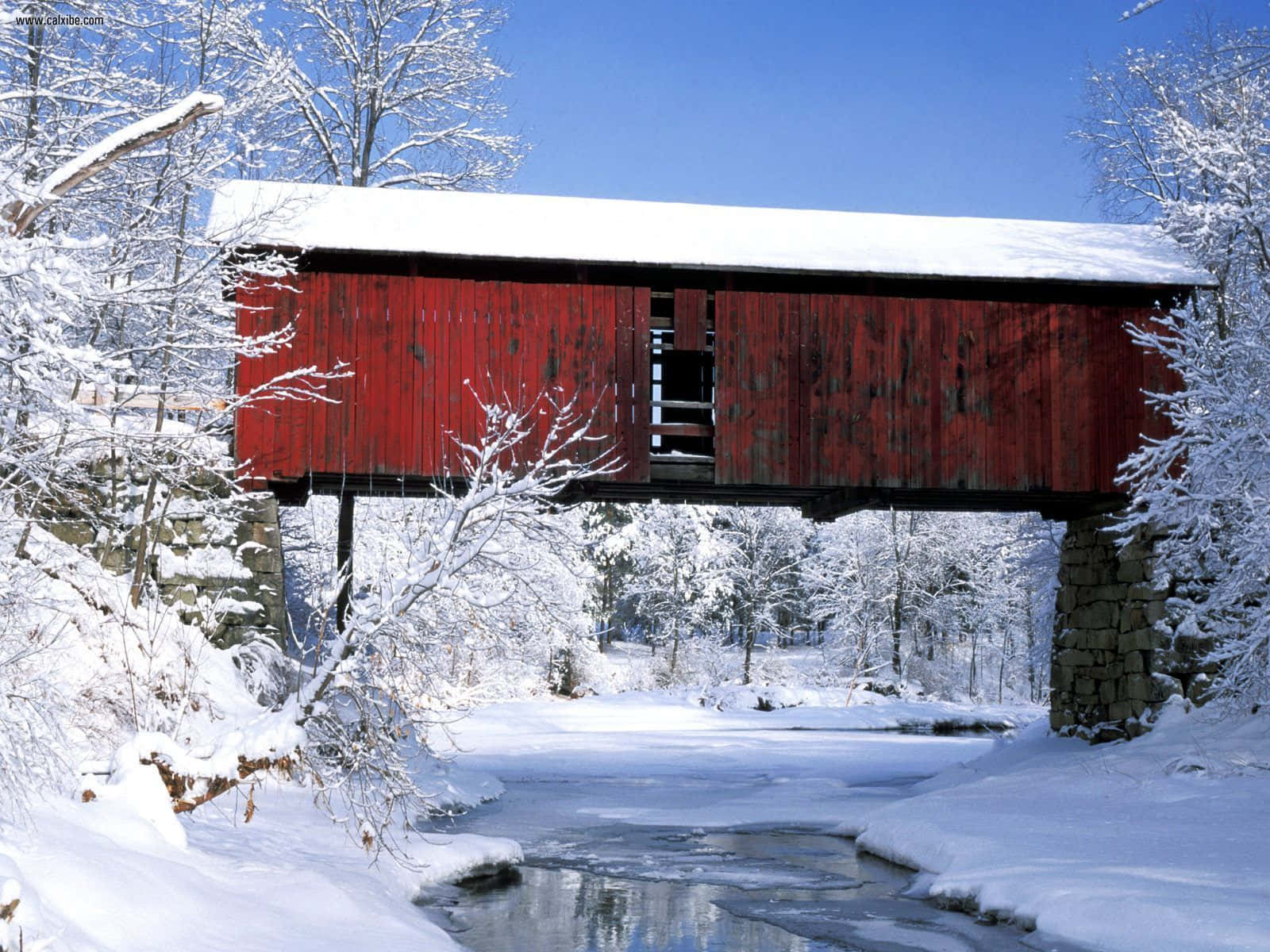 Winter Scene 桌布 壁紙 — 一座充滿活力的紅色木製有蓋橋樑被白雪覆蓋，橫跨一條部分結冰的小溪，在清澈的藍色冬日天空下。