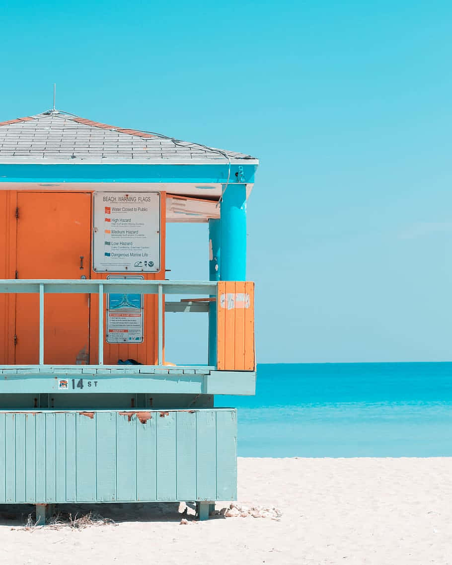Minimalist Beach 桌布 壁紙 — 一座充滿活力的藍綠色和橙色救生塔矗立在純淨的白色沙灘上，清澈的藍天下，平靜的藍綠色海水延伸至地平線。