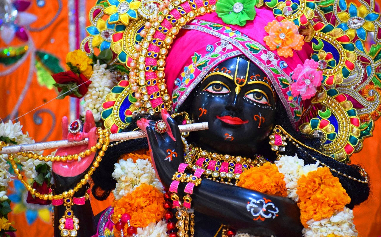 Bal Krishna 桌布 壁紙 — 一座充滿活力的黑色皮膚印度教神像，飾有粉金和色彩繽紛的花朵，在萬壽菊環繞下吹奏長笛。