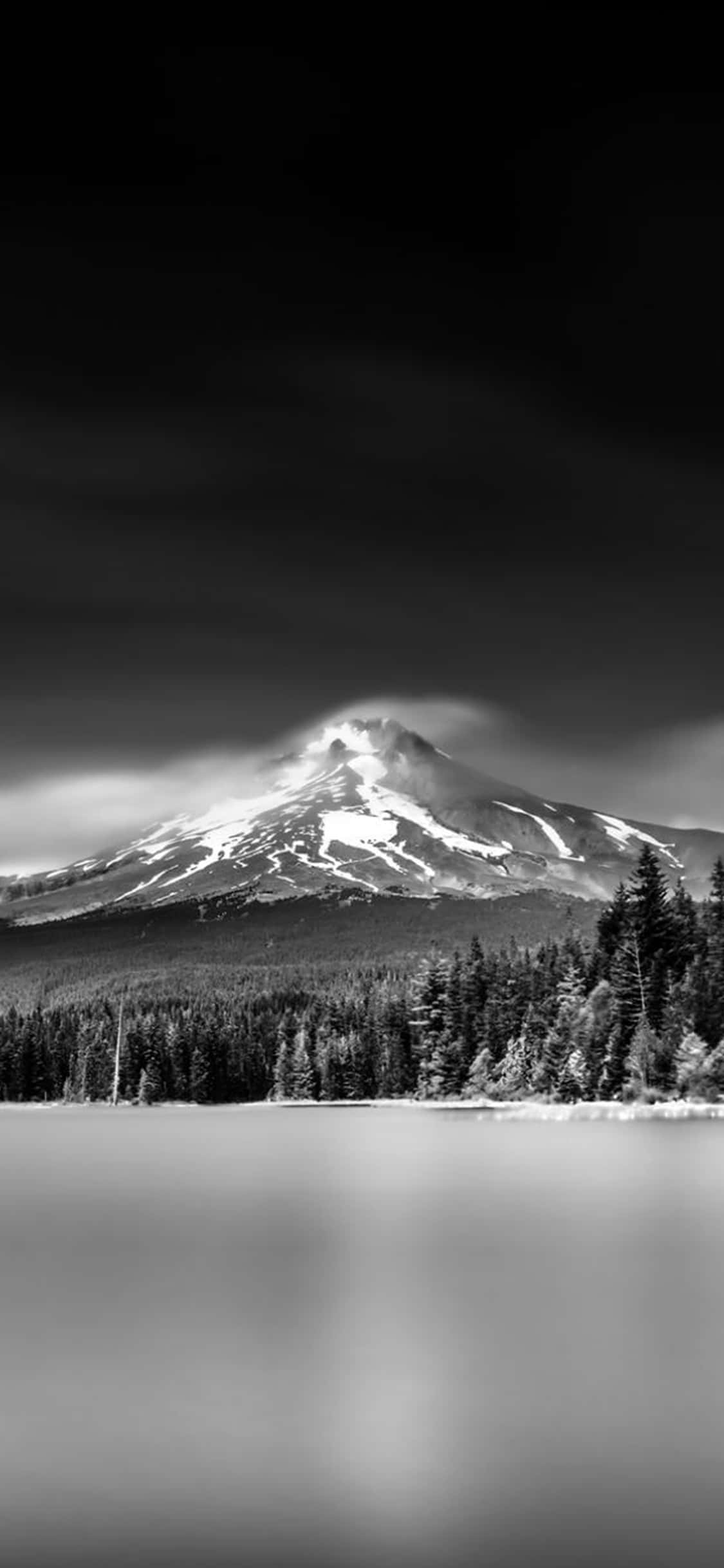 iPhone X Nature 桌布 壁紙 — 一座單色調的山峰，山頂積雪，薄霧在茂密森林環繞的平靜湖面上裊裊升起。