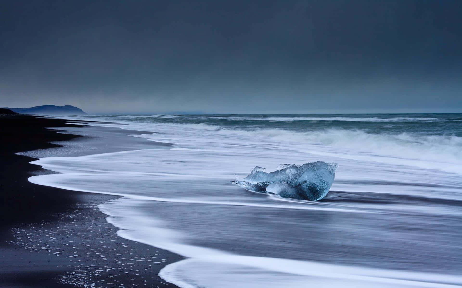Winter Beach 桌布 壁紙 — 一座孤獨的半透明藍色冰山坐落於黑色火山沙灘上，海浪在陰沉的灰色天空下拍打。